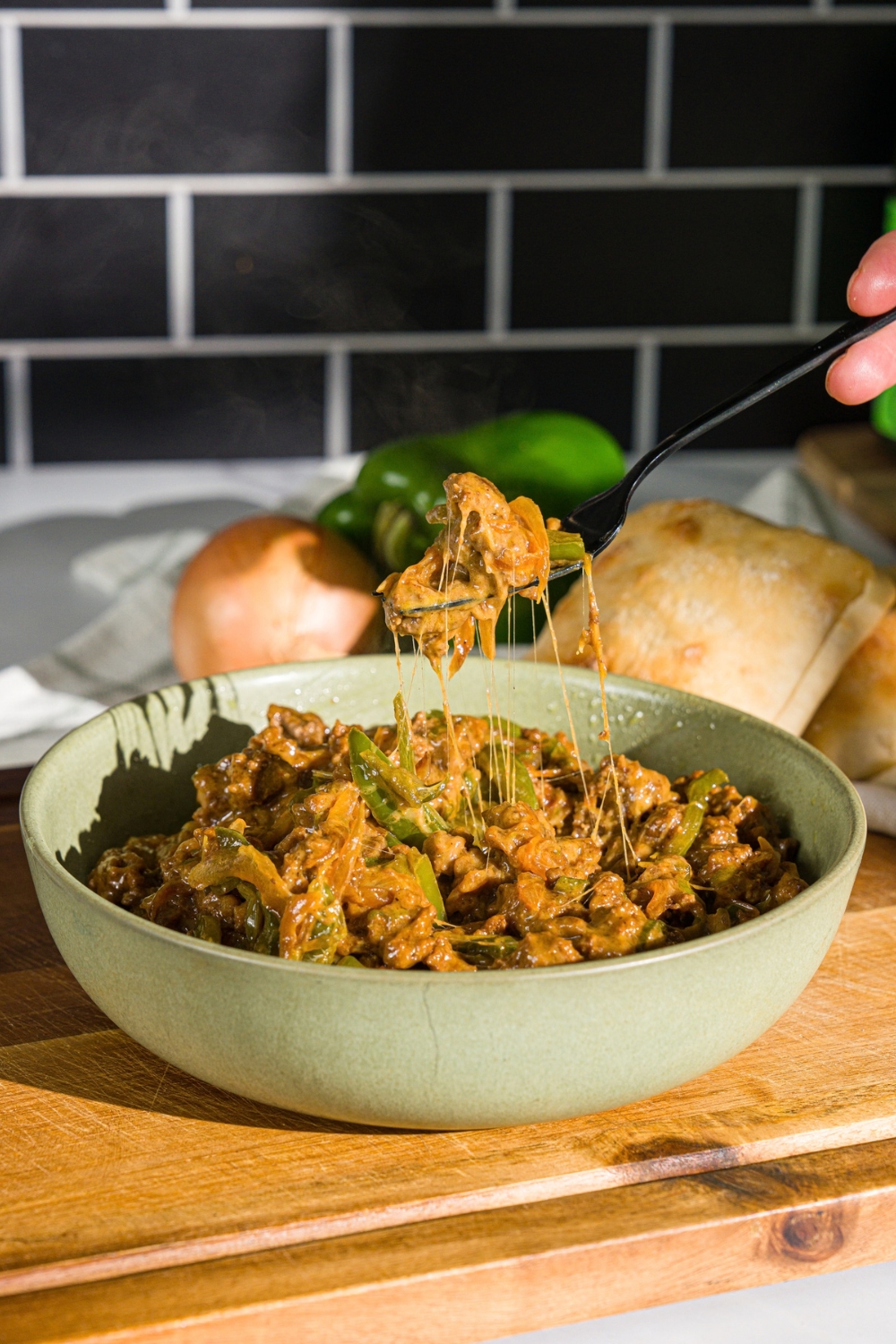 A green bowl with a philly cheesesteak bowl with ground beef, green peppers, and cheese. A fork is taking a bite from the bowl. The bowl is on a wooden board.