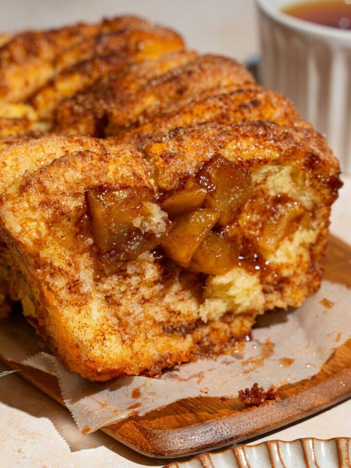 A loaf of apple pie pull apart bread on a wooden board lined with parchment paper. The first slice has been removed. The board is on a tan counter.