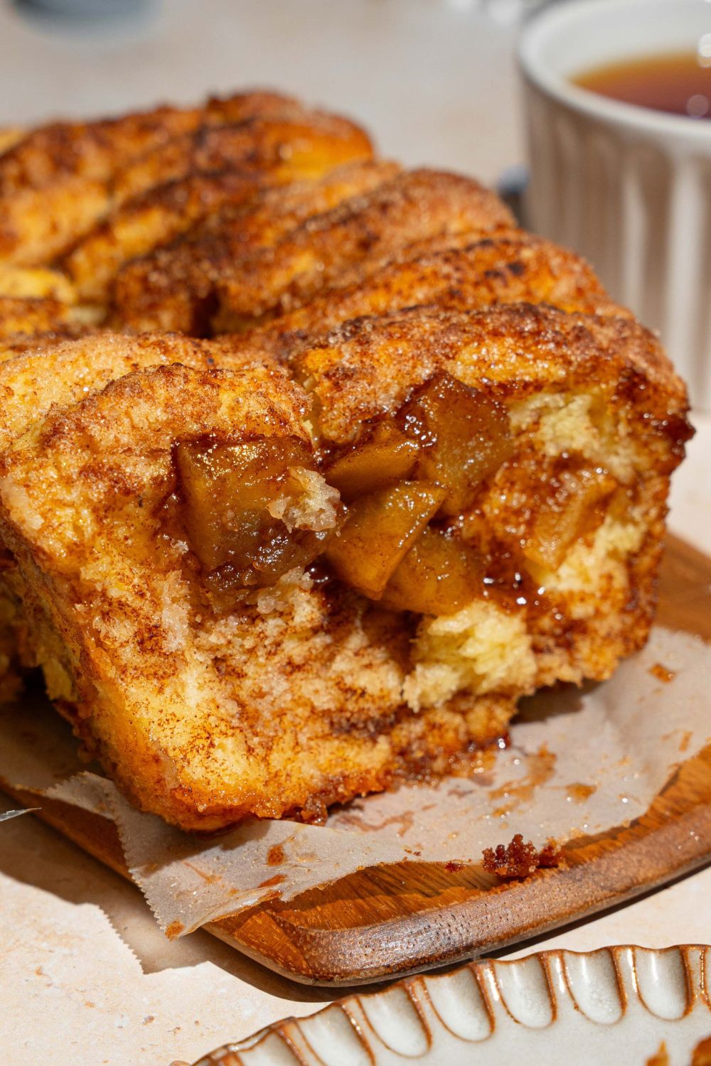 A loaf of apple pie pull apart bread on a wooden board lined with parchment paper. The first slice has been removed. The board is on a tan counter.