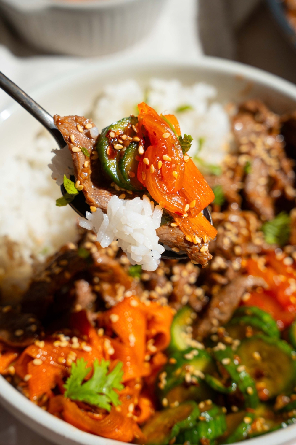 A close up of a fork with a bite of beef bulgogi including rice, beef, and cucumber carrot salad garnished with sesame seeds. There is a beef bulgogi bowl blurred in the background.