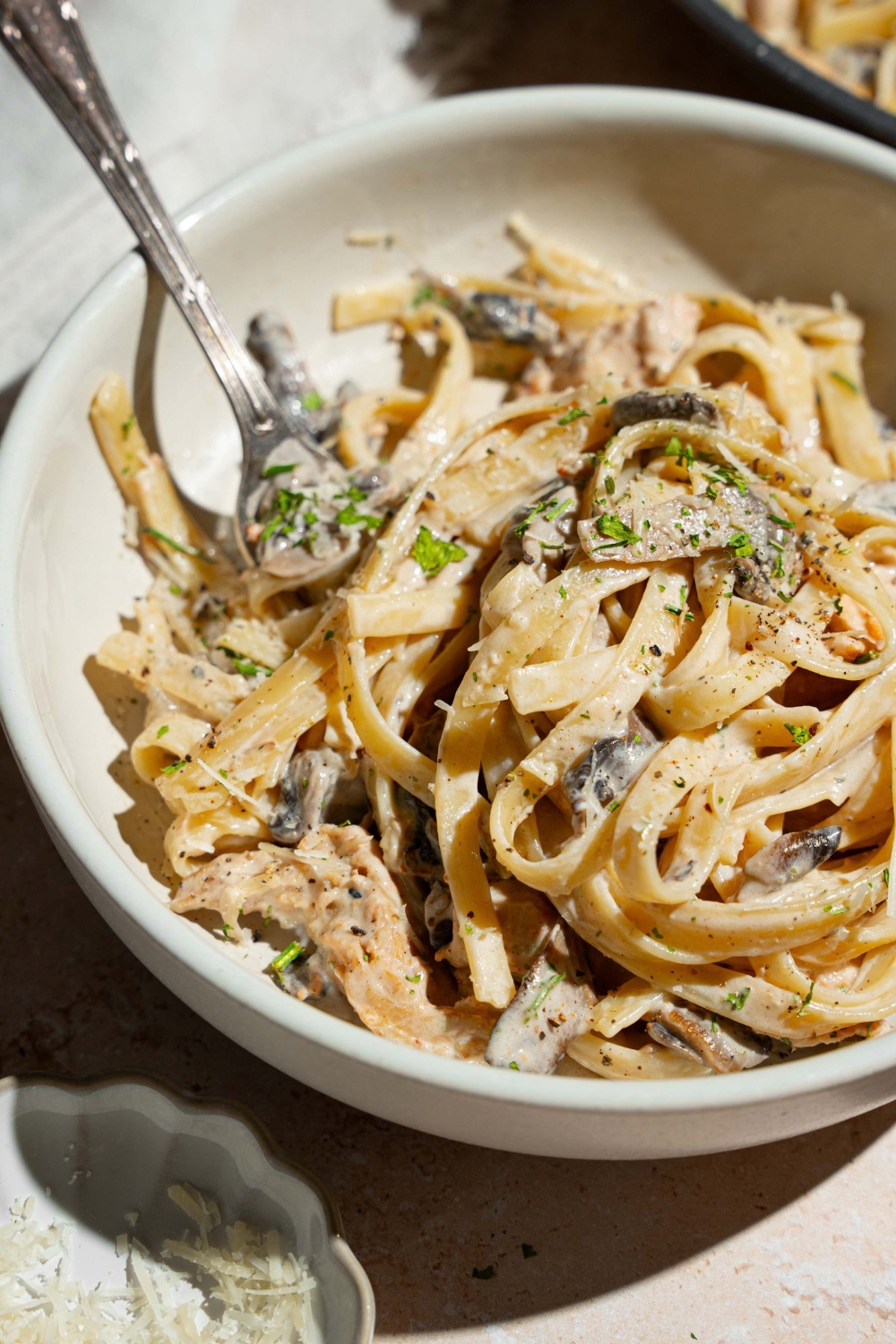 A white plate with chicken mushroom fettuccine alfredo garnished with pepper and parmesan. There is a fork in the pasta.