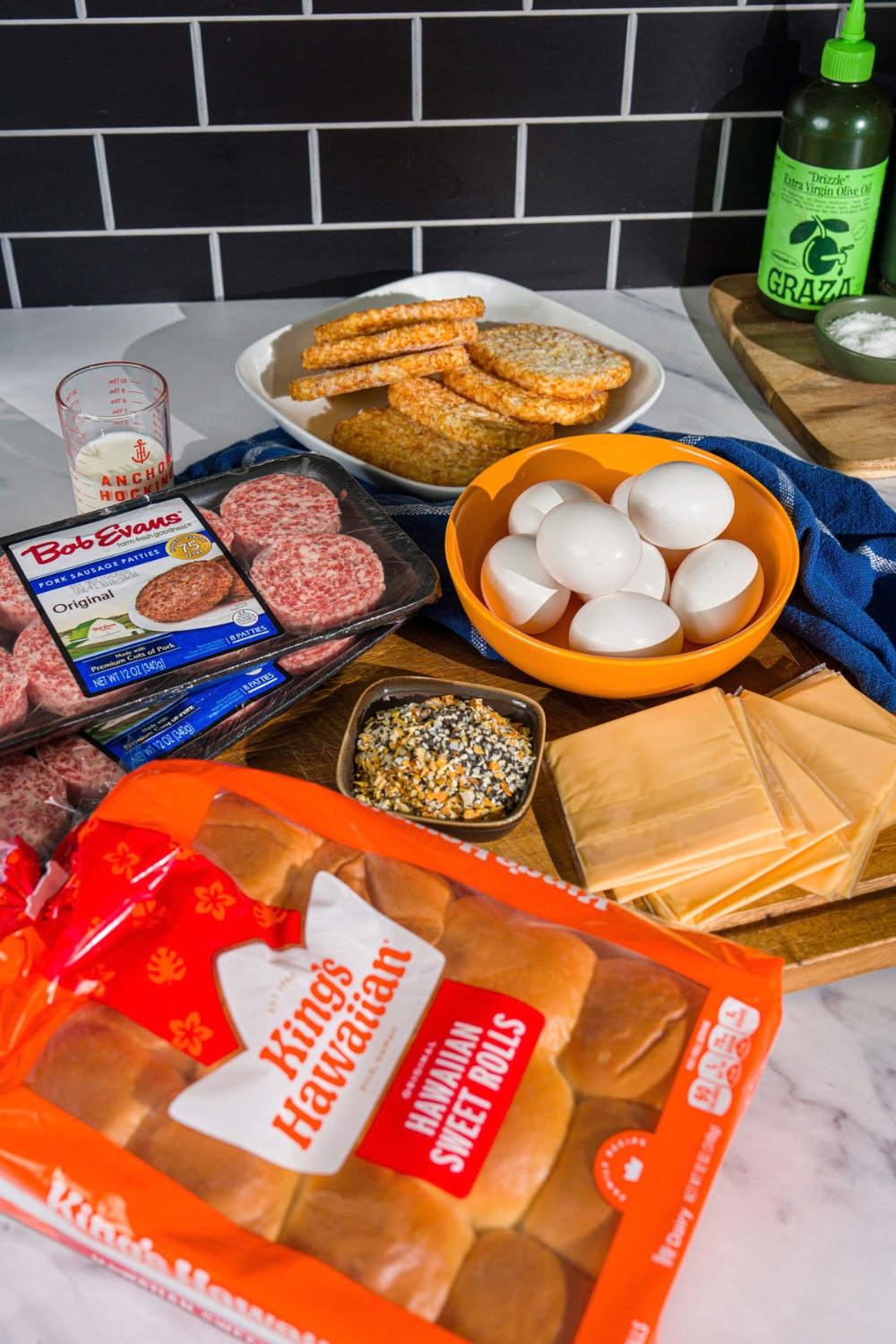 A wooden board with ingredients to make breakfast sliders including eggs, hawaiian rolls, cheese slices, sausage patties, hashbrowns, milk, and seasonings.