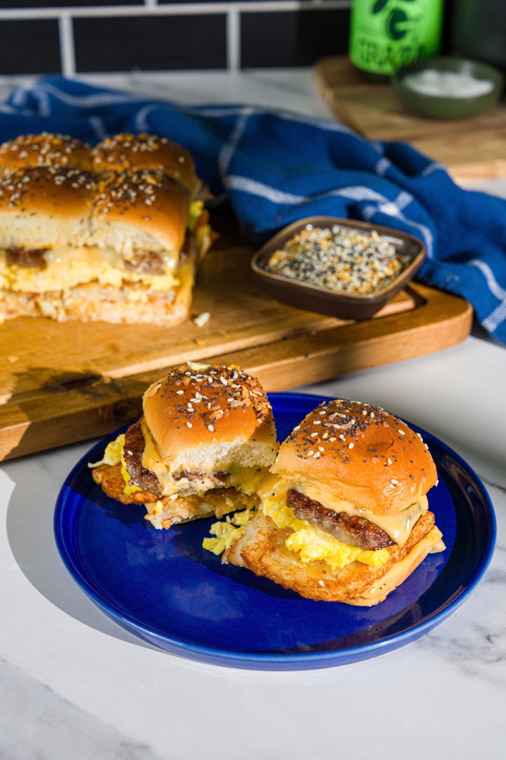 A blue plate with two breakfast sliders made with sausage, hashbrowns, scrambled eggs, and cheese on a seasoned hawaiian roll. The plate is on a marble counter with a wooden board with sliders.