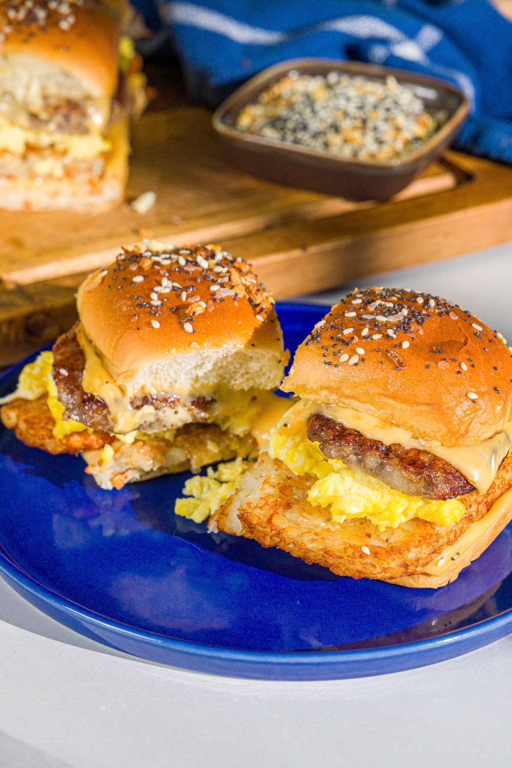 A blue plate with two breakfast sliders made with sausage, hashbrowns, scrambled eggs, and cheese on a seasoned hawaiian roll. The plate is on a marble counter with a wooden board with sliders.