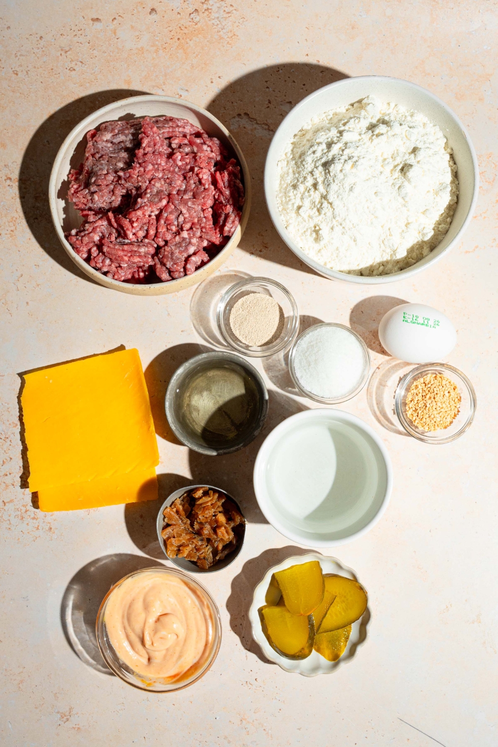 A tan counter with ingredients to make burger stuffed buns including ground beef, flour, yeast, sugar, egg, cheese, pickles, burger sauce, and more.