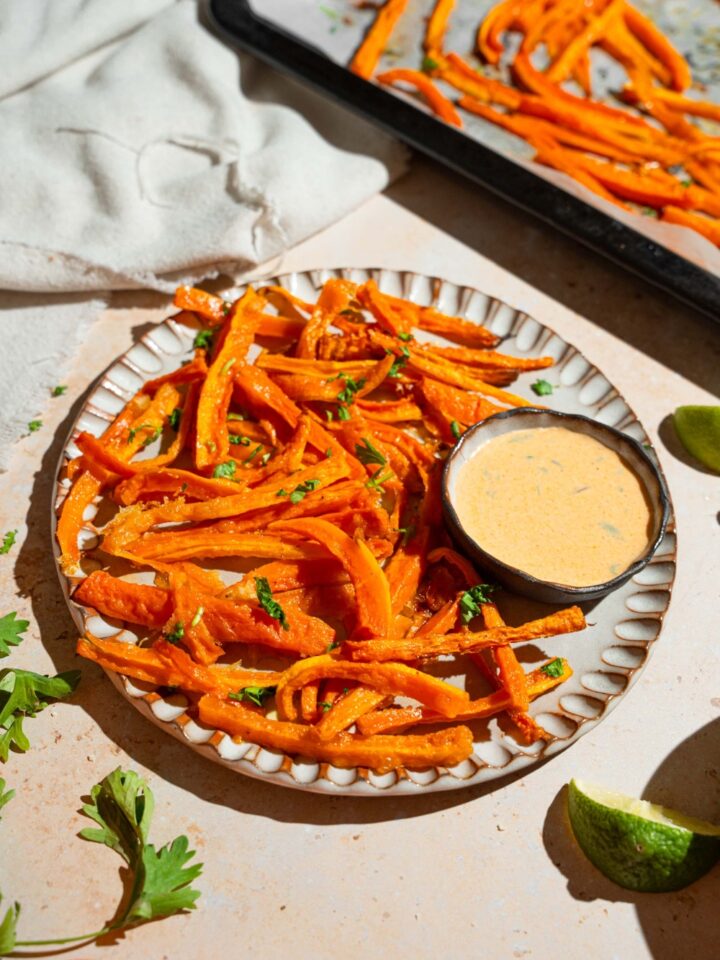 A plate of carrot fries garnished with fresh parsley and served with a bowl fo dipping sauce. The plate is on a tan counter with a baking sheet of fries and a white cloth napkin.