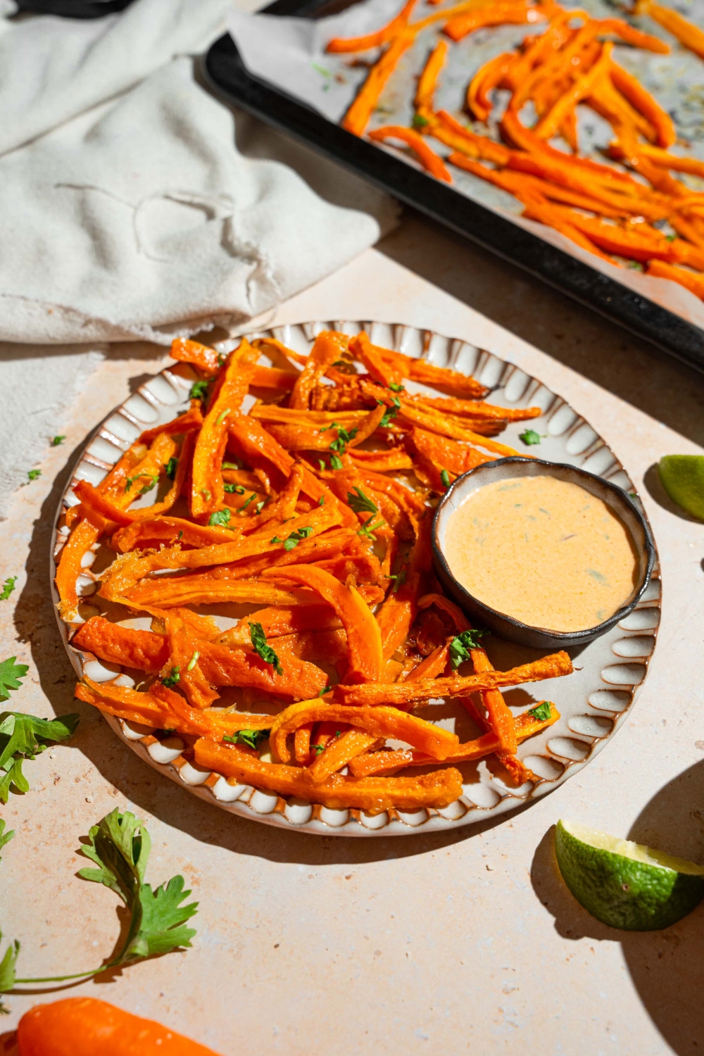 A plate of carrot fries garnished with fresh parsley and served with a bowl fo dipping sauce. The plate is on a tan counter with a baking sheet of fries and a white cloth napkin.