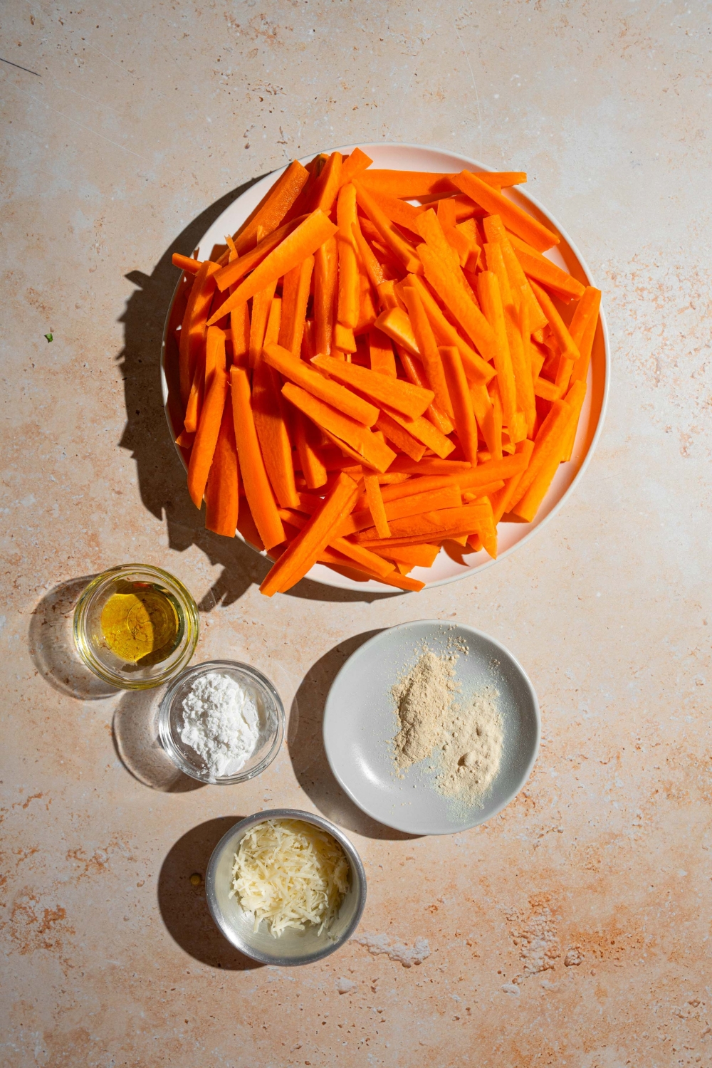 A tan counter with several bowls containing ingredients to make carrot fries including sliced carrots, cornstarch, cheese, oil, and more.