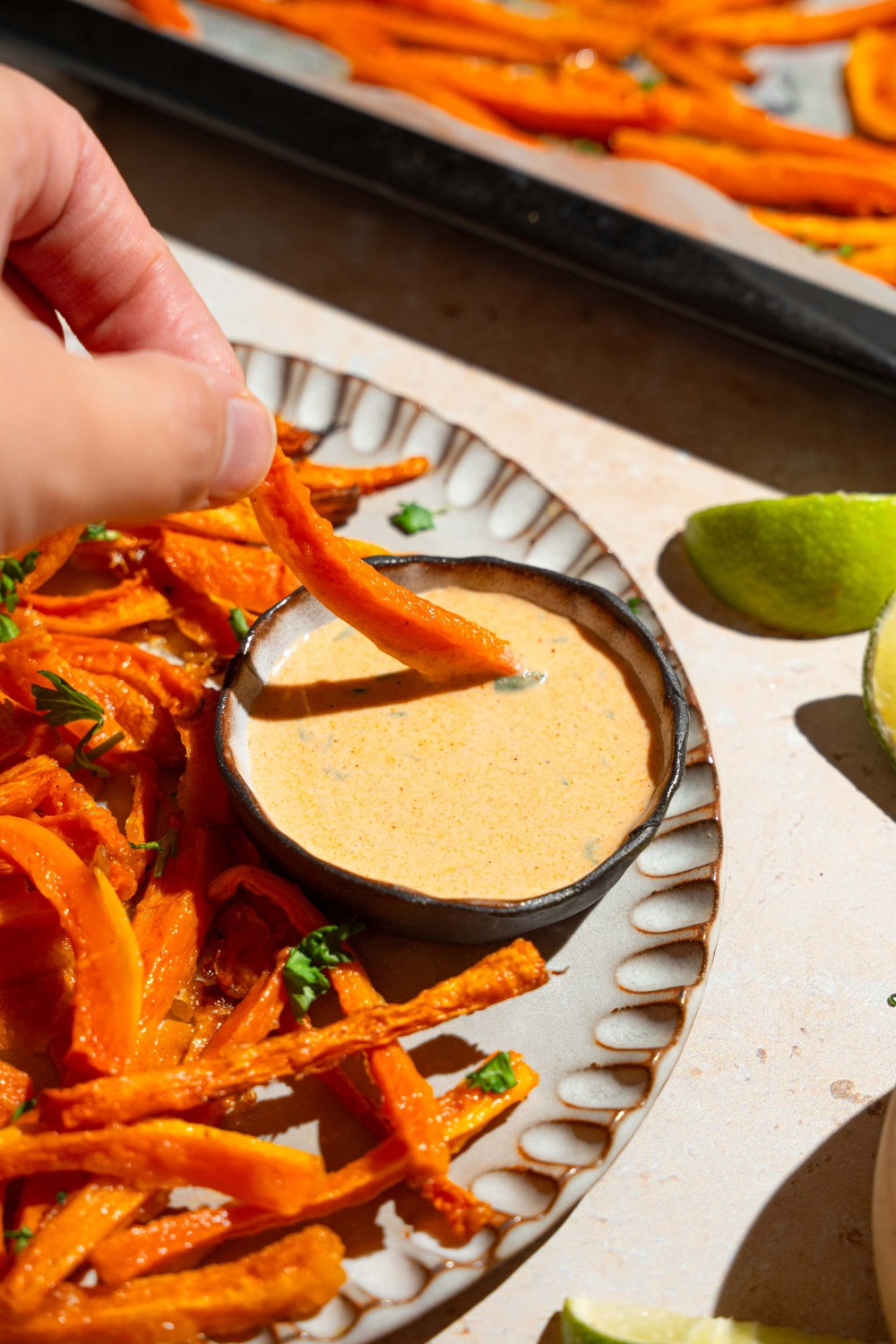 A plate of carrot fries garnished with fresh parsley and served with a bowl fo dipping sauce. A fry is being dipped into the sauce. The plate is on a tan counter with a baking sheet of fries and a white cloth napkin.