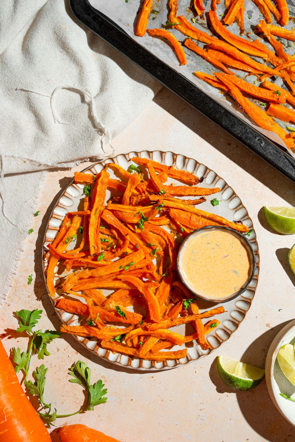 A plate of carrot fries garnished with fresh parsley and served with a bowl fo dipping sauce. The plate is on a tan counter with a baking sheet of fries and a white cloth napkin.