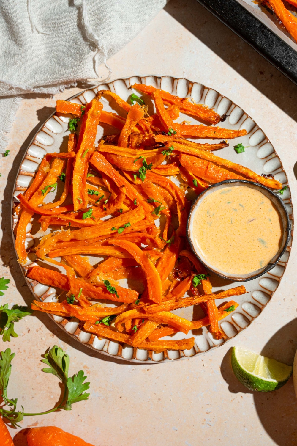 A plate of carrot fries garnished with fresh parsley and served with a bowl fo dipping sauce. The plate is on a tan counter with a baking sheet of fries and a white cloth napkin.