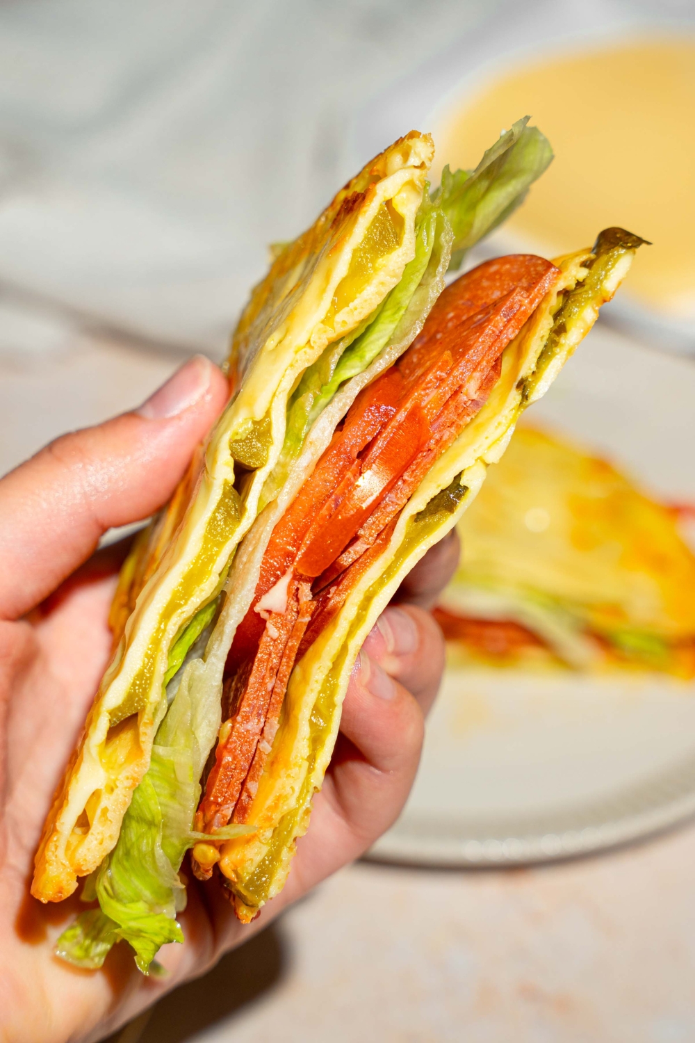 A close up of a hand holding a sandwich with pickle bread, lettuce, and tomato. There is a plate with the rest of the sandwich blurred in the background.