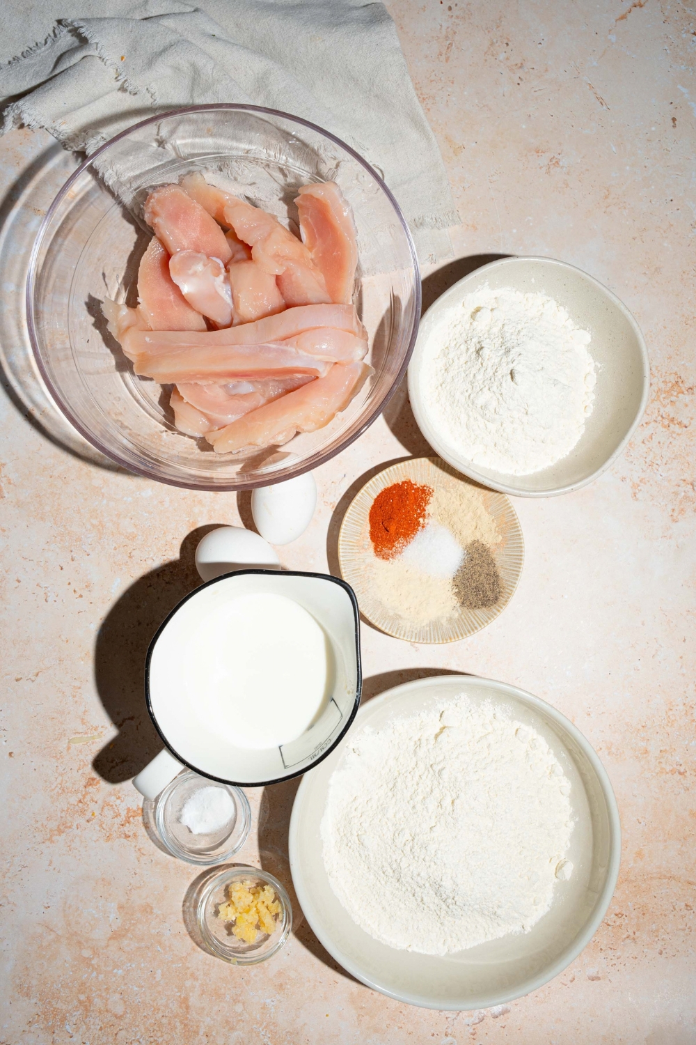 A tan counter with several bowls containing ingredients to make copycat Chick-Fil-A tenders including chicken, buttermilk, flour, eggs, garlic, and seasonings.