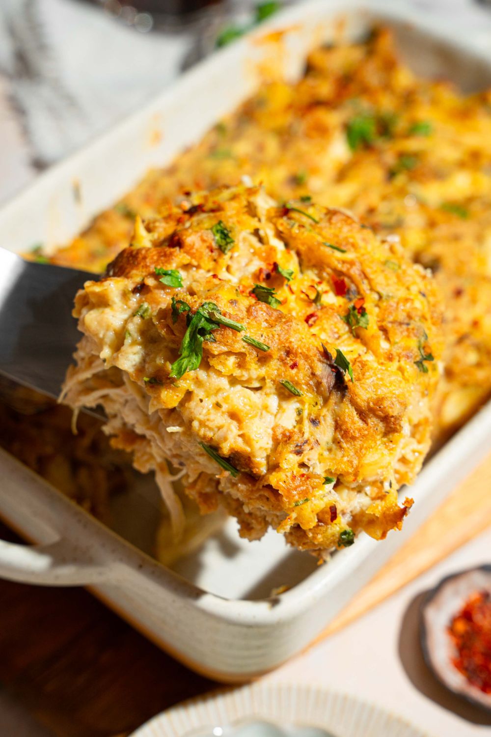 A close up of a spatula with a piece of Dolly Parton's chicken and stuffing casserole garnished with fresh parsley. There is a baking dish of casserole blurred in the background.