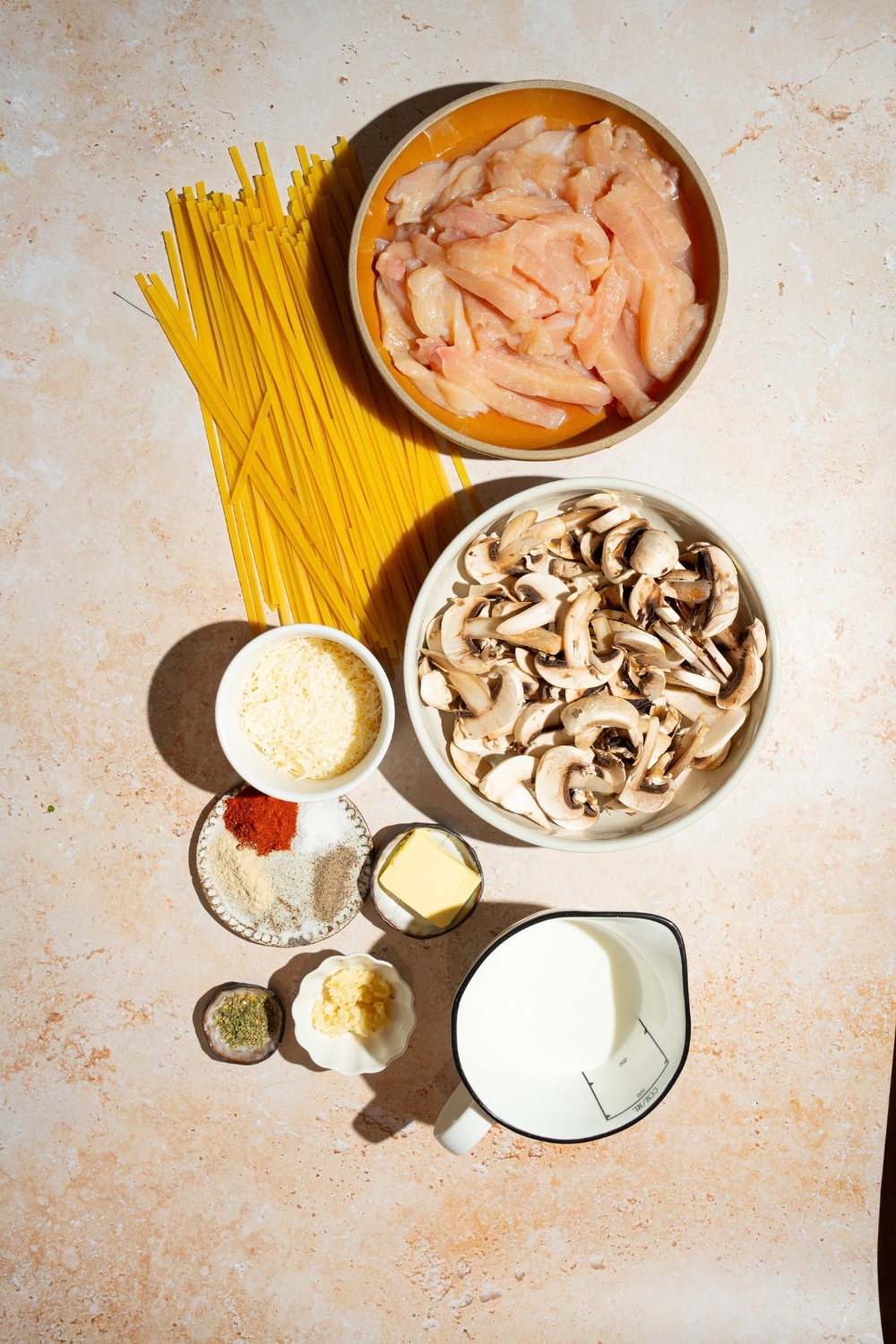 A tan counter with ingredients to make chicken mushroom alfredo including chicken, mushrooms, fettuccine pasta, parmesan cheese, garlic, butter, heavy cream, and seasonings.