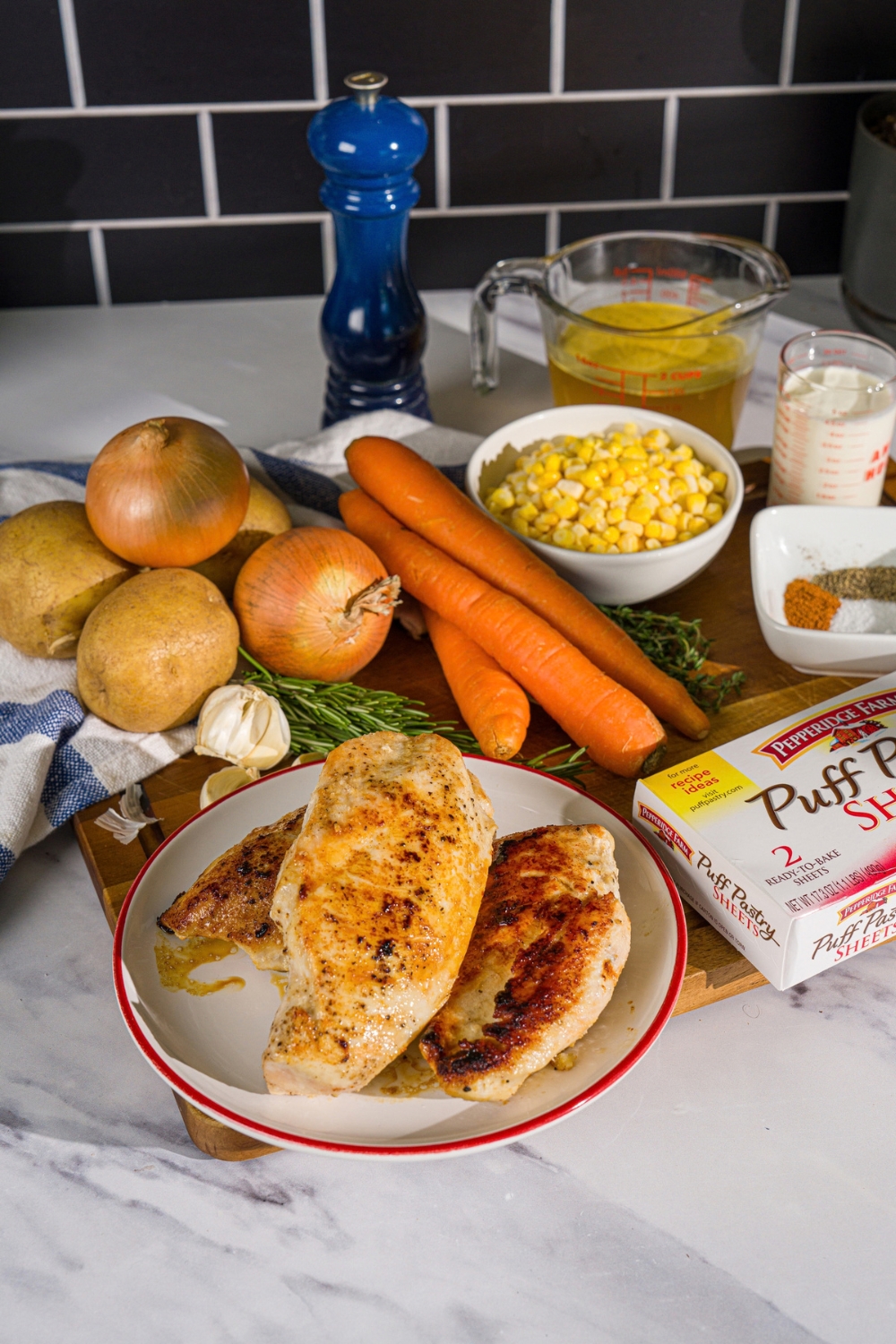 A wooden board with ingredients to make chicken pot pie hand pies including grilled chicken breasts, corn, carrots, potatoes, onion, puff pastry, chick stock, and more.
