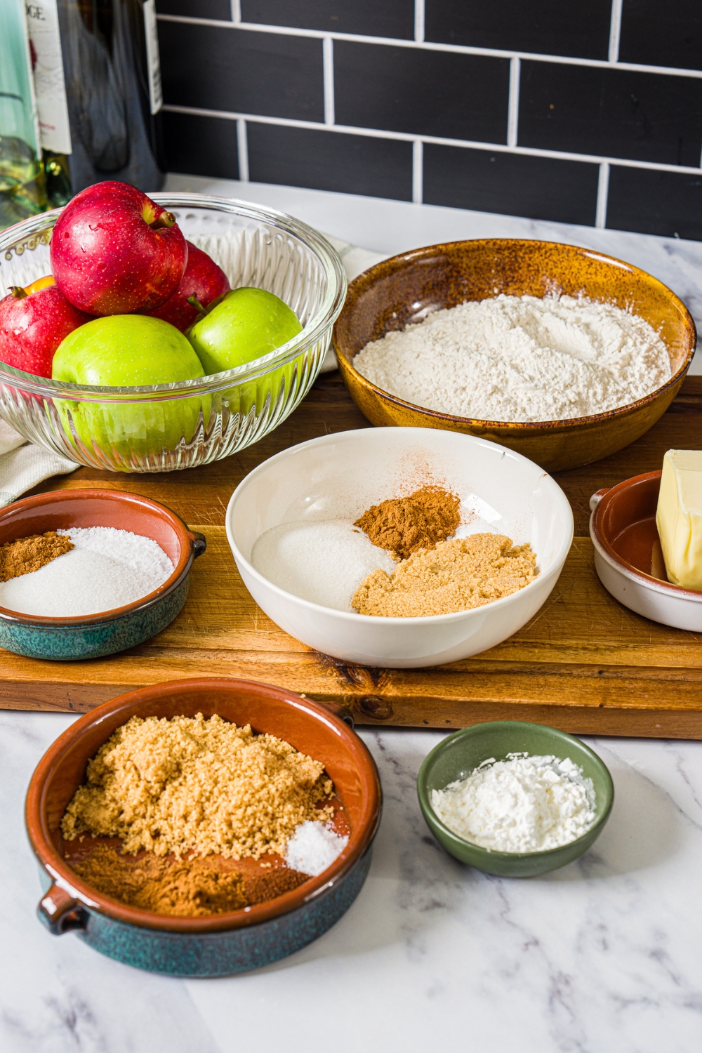 A wooden board with ingredients to make cinnamon roll apple pie including flour, sugar, brown sugar, cinnamon, apples, butter, cornstarch, and seasonings.