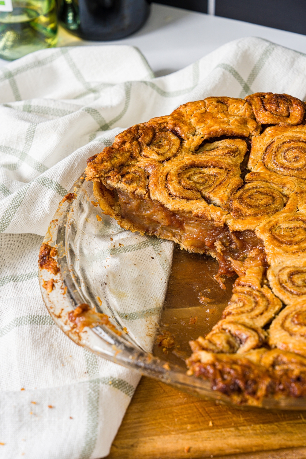 A glass pie dish with baked cinnamon roll apple pie with slices removed. The dish is on a wooden board with a white cloth napkin.