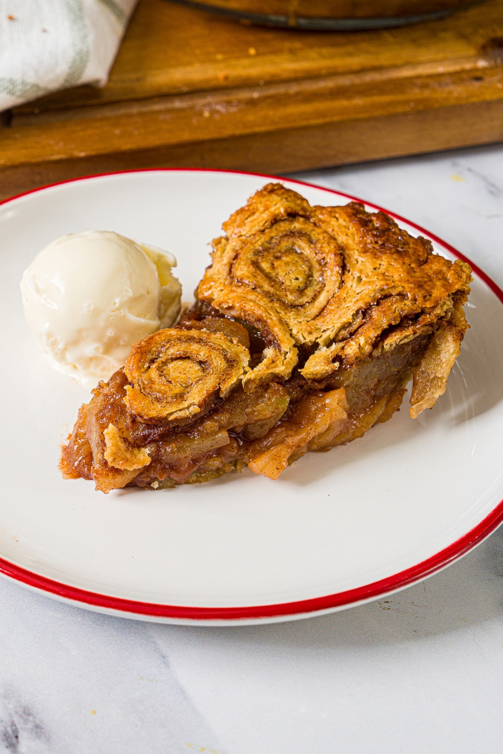 A white plate with a slice of apple pie with cinnamon roll crust served with a scoop of vanilla ice cream. The plate is on a marble counter.