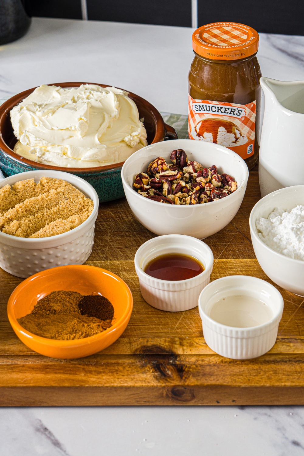 A bowl of cream cheese, a bowl of pecans, a bowl of brown sugar, a bowl cinnamon, and a bowl of powdered sugar all on a wood board.