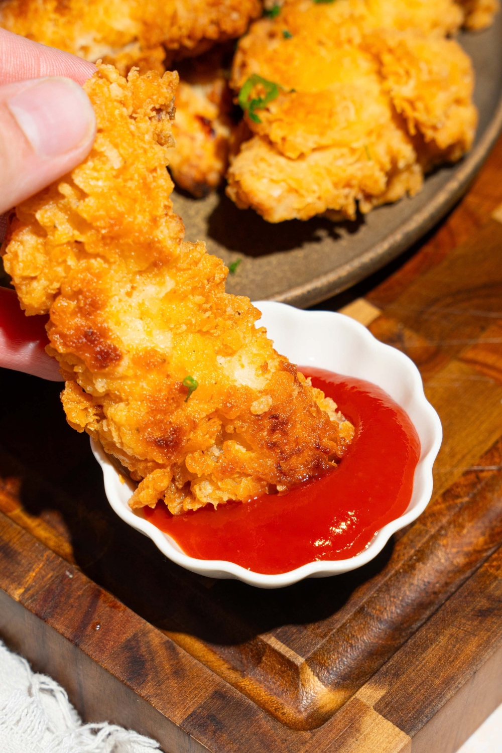 A hand dipping a copycat Chick-Fil-A tender into a small bowl of dipping sauce. The bowl is on a wooden board with a plate of tenders.