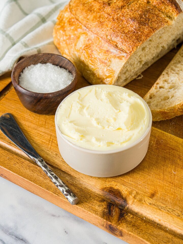 A small bowl of cultured butter on a wooden board with a butter knife, small bowl of salt, and loaf of bread.