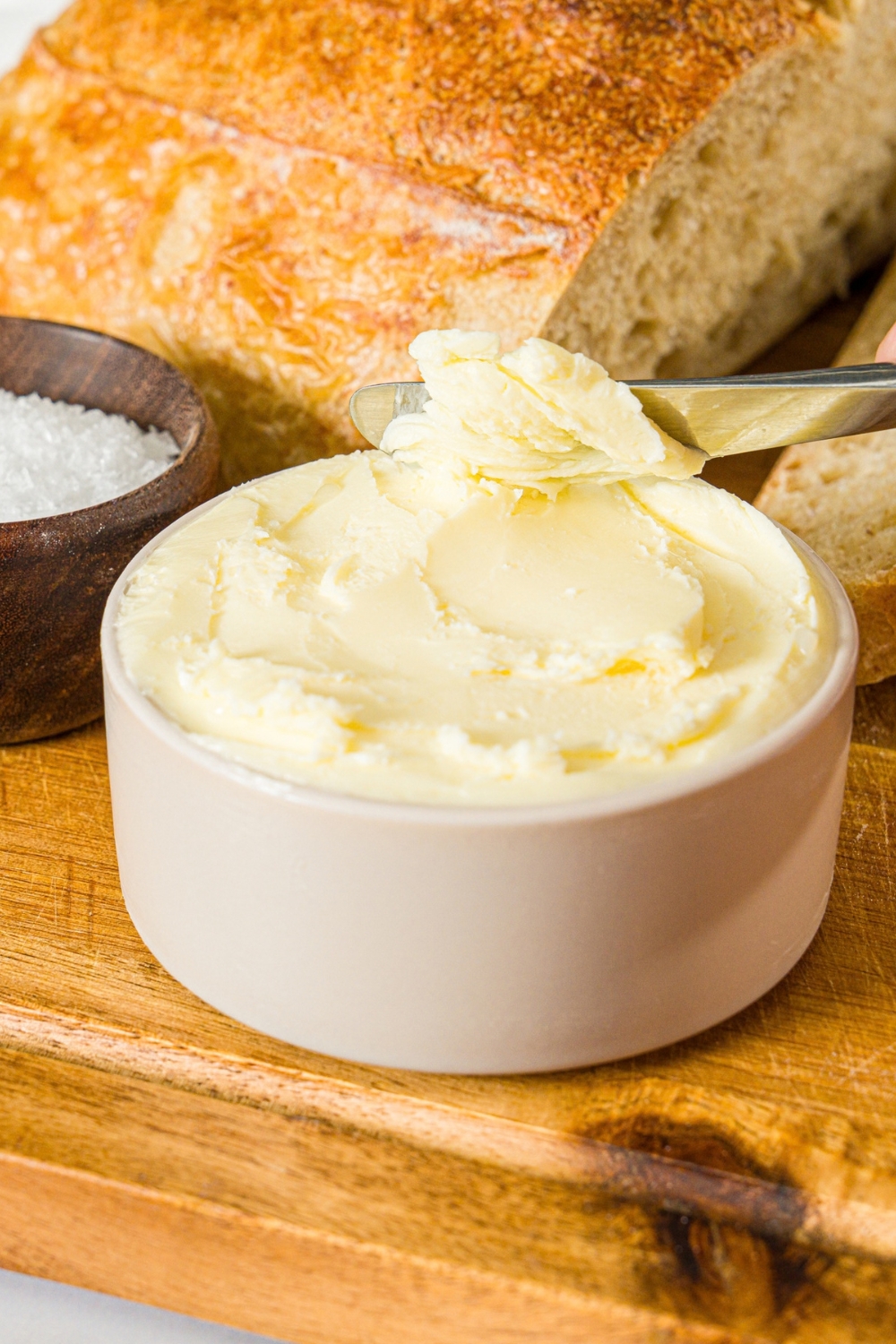 A small bowl of cultured butter. A butter knife is dipped into the butter. The bowl is on a wooden board with a bowl of salt and loaf of bread.