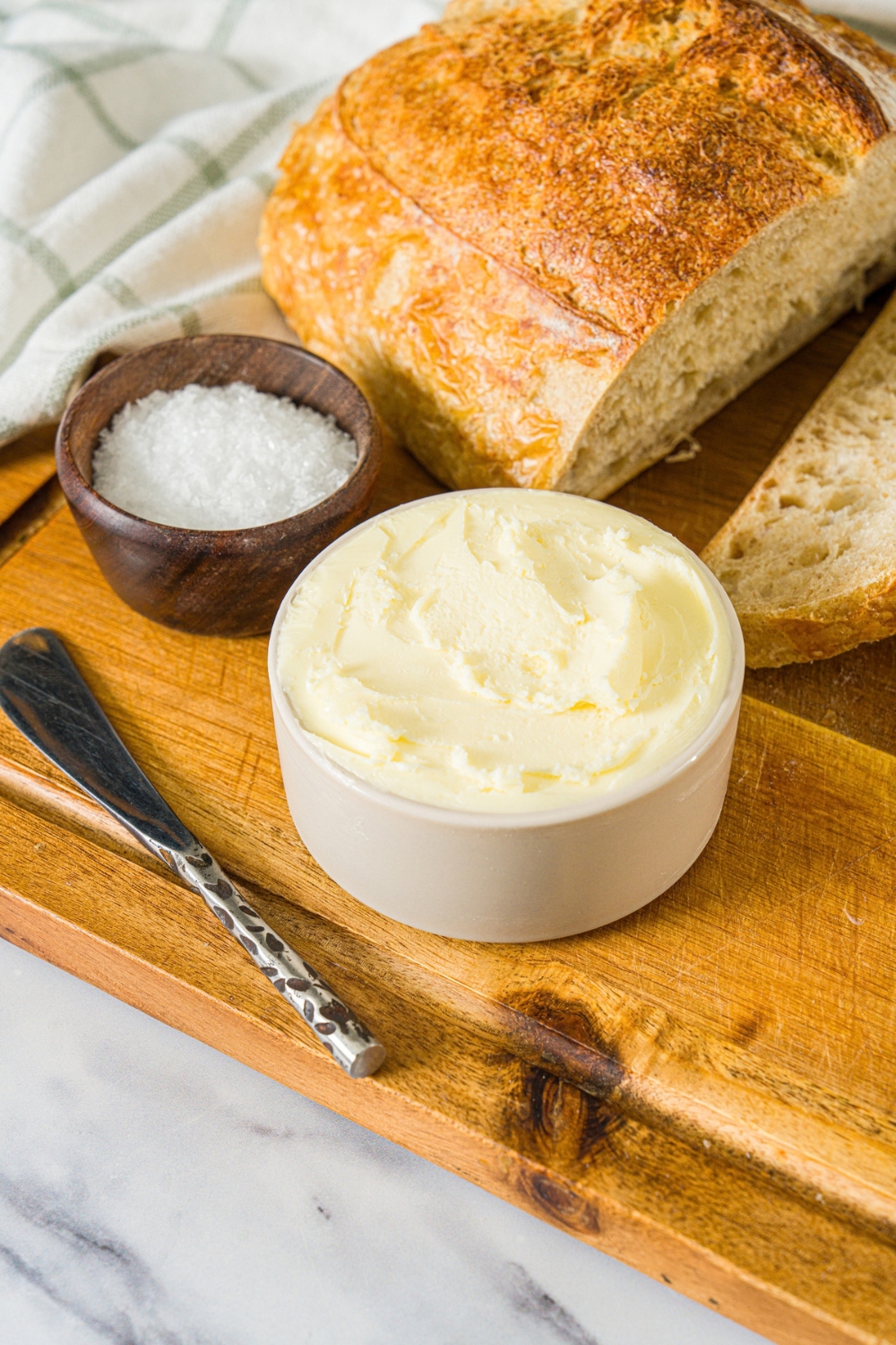 A small bowl of cultured butter on a wooden board with a butter knife, small bowl of salt, and loaf of bread.