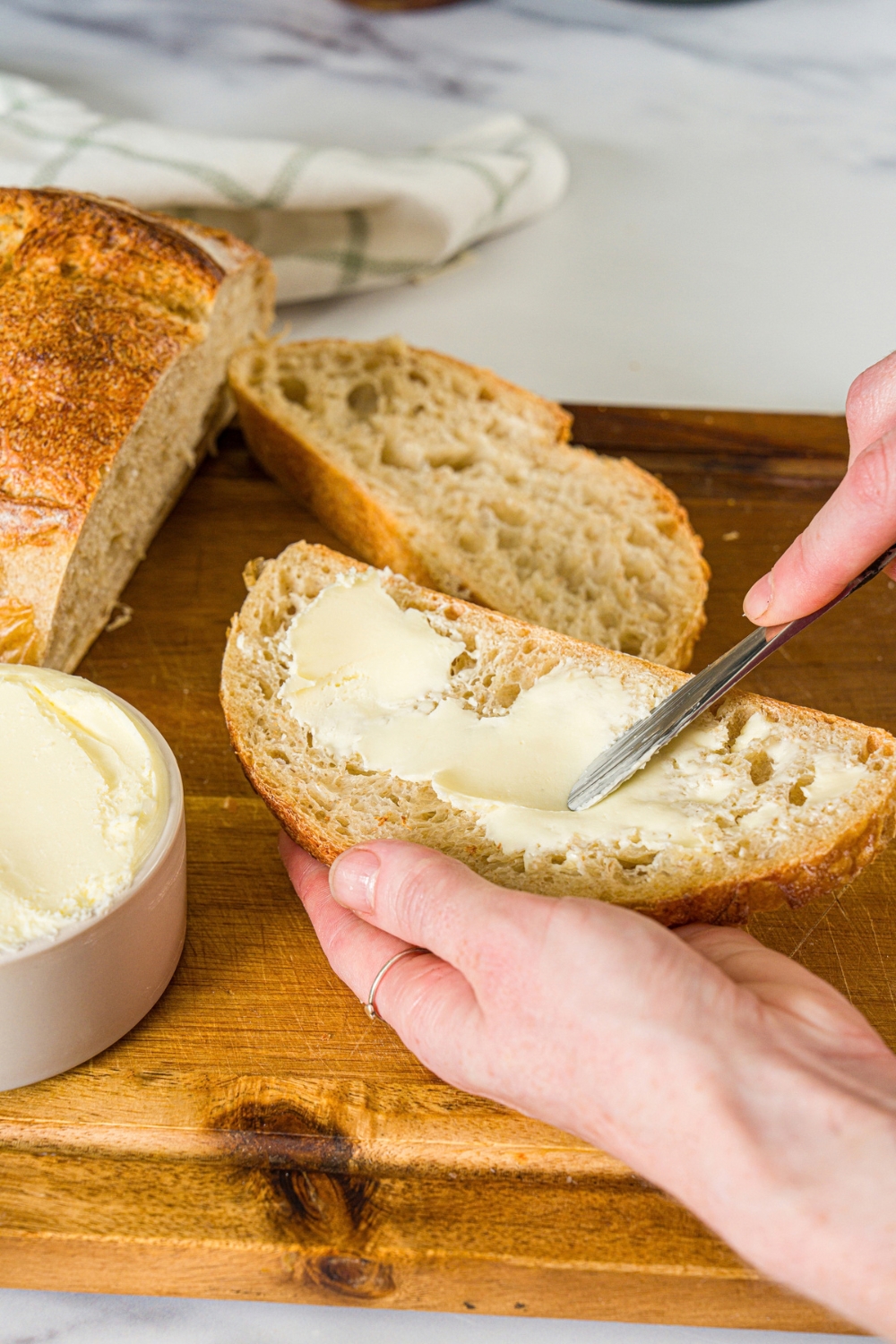 A butter knife spreading cultured butter on a slice of bread. There is a wooden board with a bowl of butter and loaf of bread in the background.