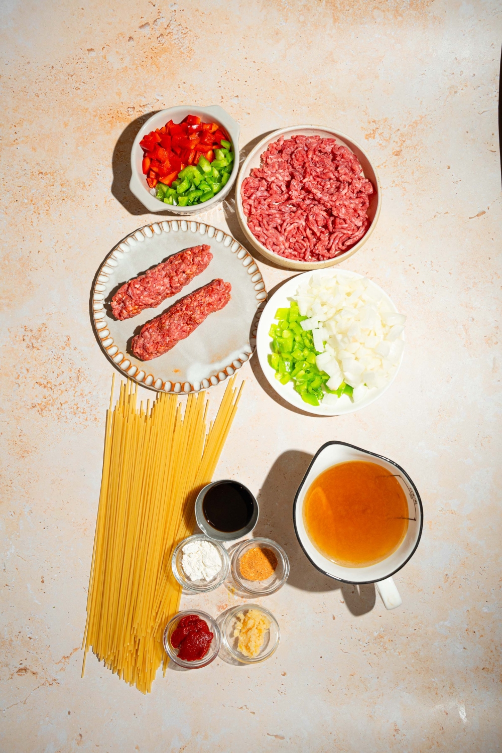 A tan counter with several bowls containing ingredients to make dirty spaghetti including diced peppers, ground beef, Italian sausage, celery, onion, stock, spaghetti, and more.