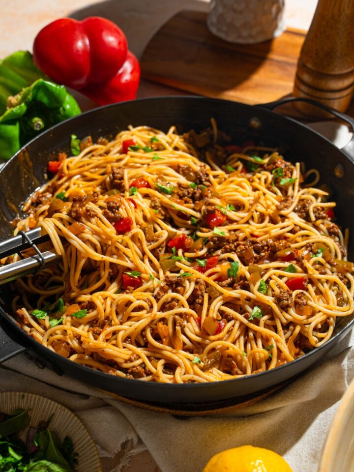 A skillet with dirty spaghetti tossed with diced peppers, celery, onion, ground beef, and Italian sausage garnished with fresh parsley. A pair of tongs is pulling some spaghetti from the skillet. The skillet is on a tan counter with peppers and small bowl of garnishes.