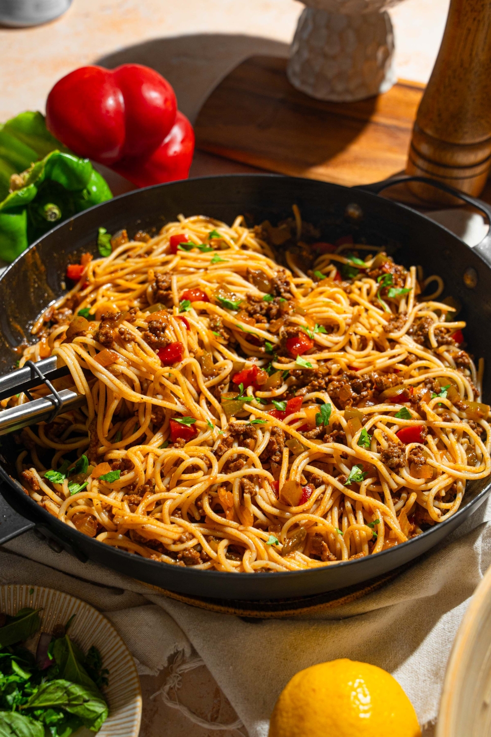 A skillet with dirty spaghetti tossed with diced peppers, celery, onion, ground beef, and Italian sausage garnished with fresh parsley. A pair of tongs is pulling some spaghetti from the skillet. The skillet is on a tan counter with peppers and small bowl of garnishes.