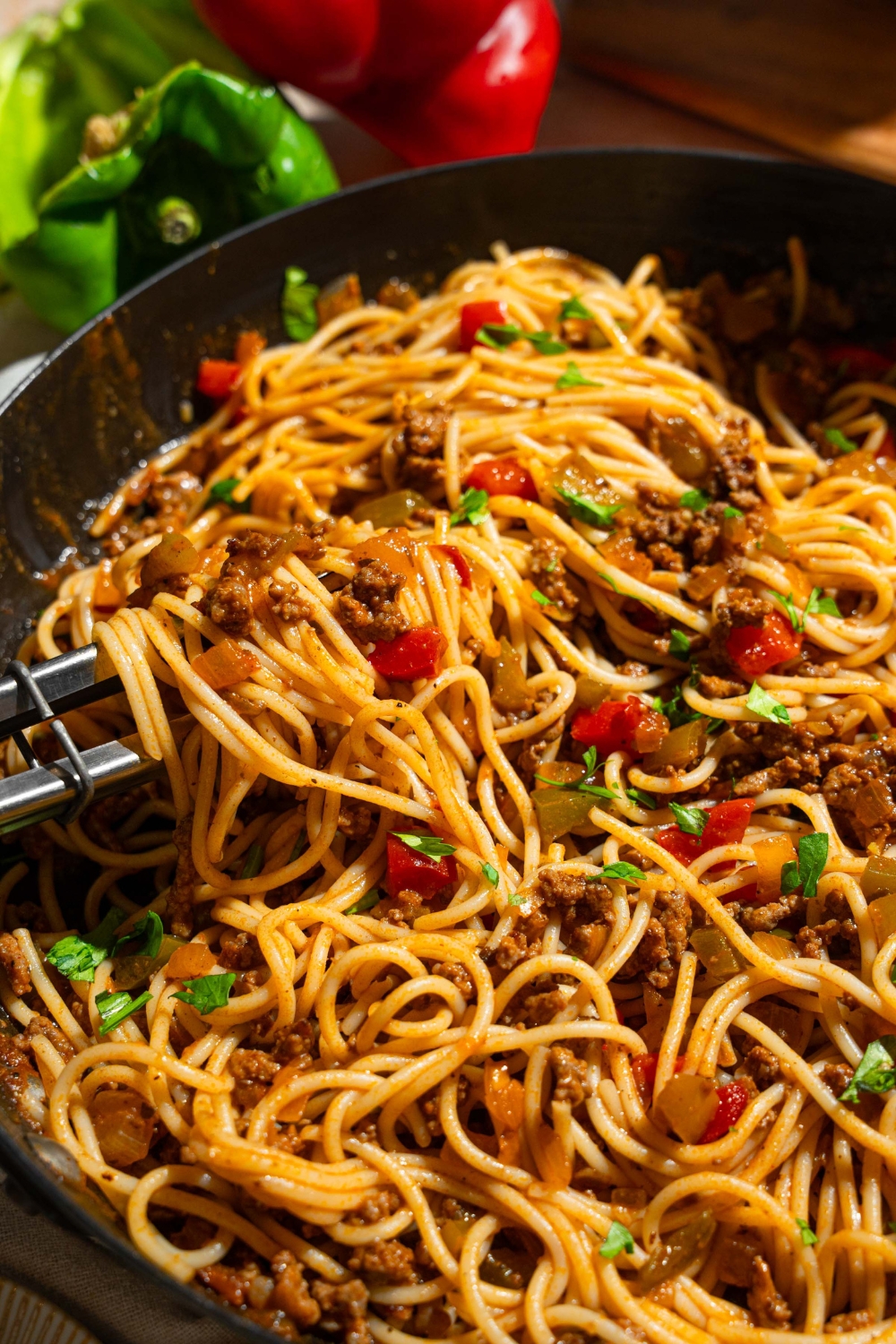 A skillet with dirty spaghetti tossed with diced peppers, celery, onion, ground beef, and Italian sausage garnished with fresh parsley. A pair of tongs is pulling some spaghetti from the skillet.