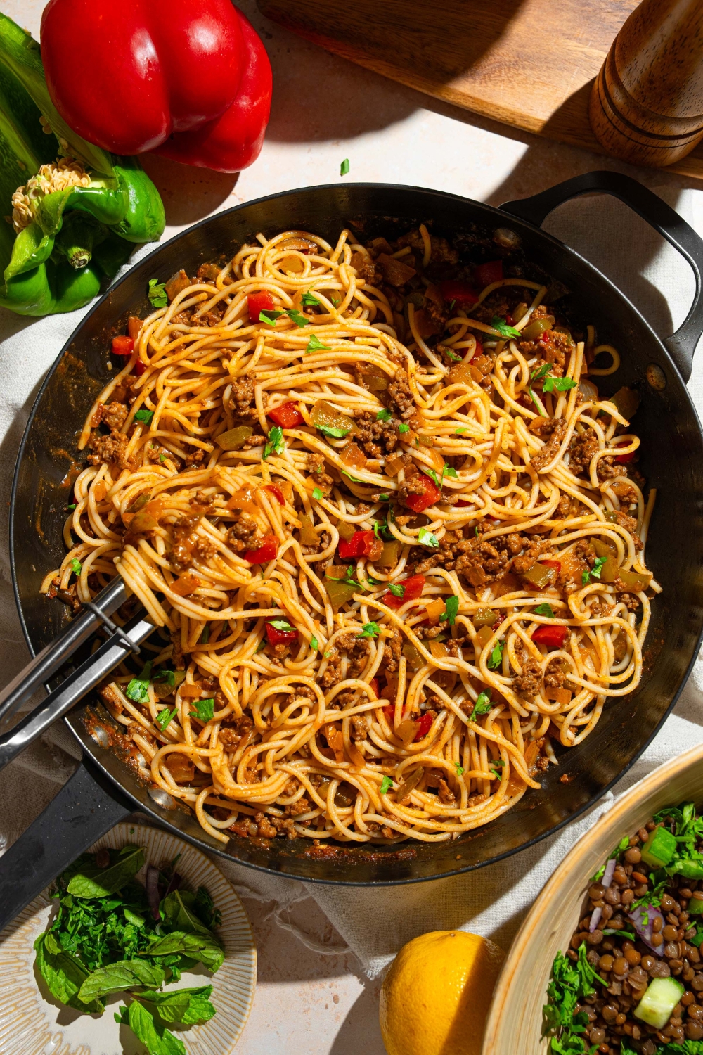 A skillet with dirty spaghetti tossed with diced peppers, celery, onion, ground beef, and Italian sausage garnished with fresh parsley. There is a pair of tongs in the spaghetti. The skillet is on a tan counter with a plate of garnishes and peppers.