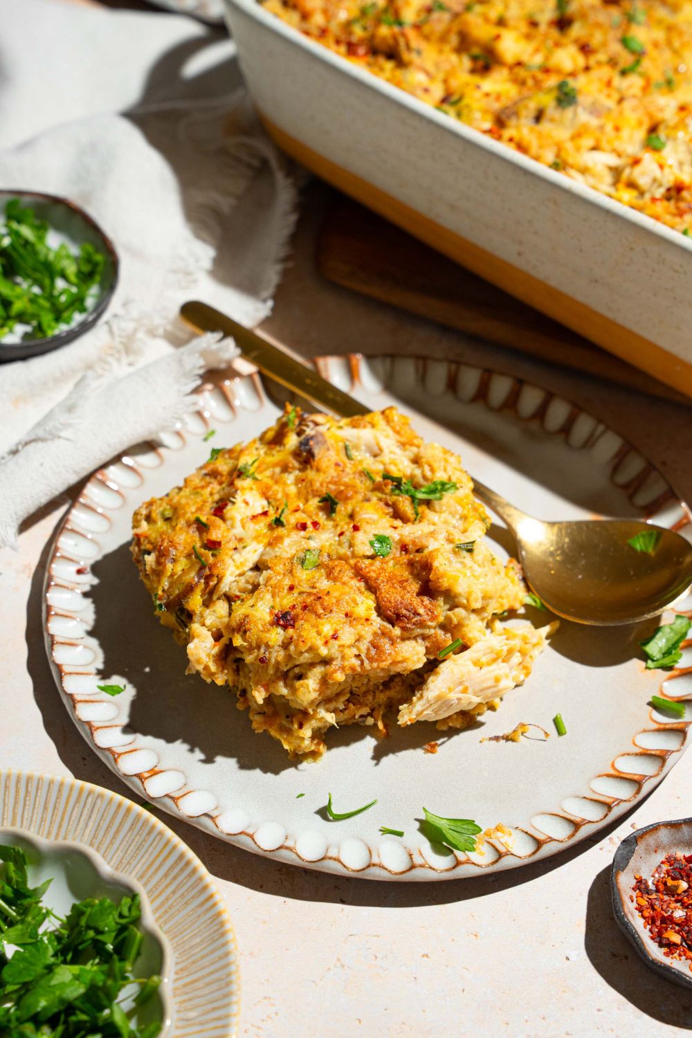 A white plate with a slice of Dolly Parton's chicken and stuffing casserole garnished with fresh parsley. There is a spoon on the plarte. The plate is on a tan counter with a baking dish of casserole and small bowls of garnishes.