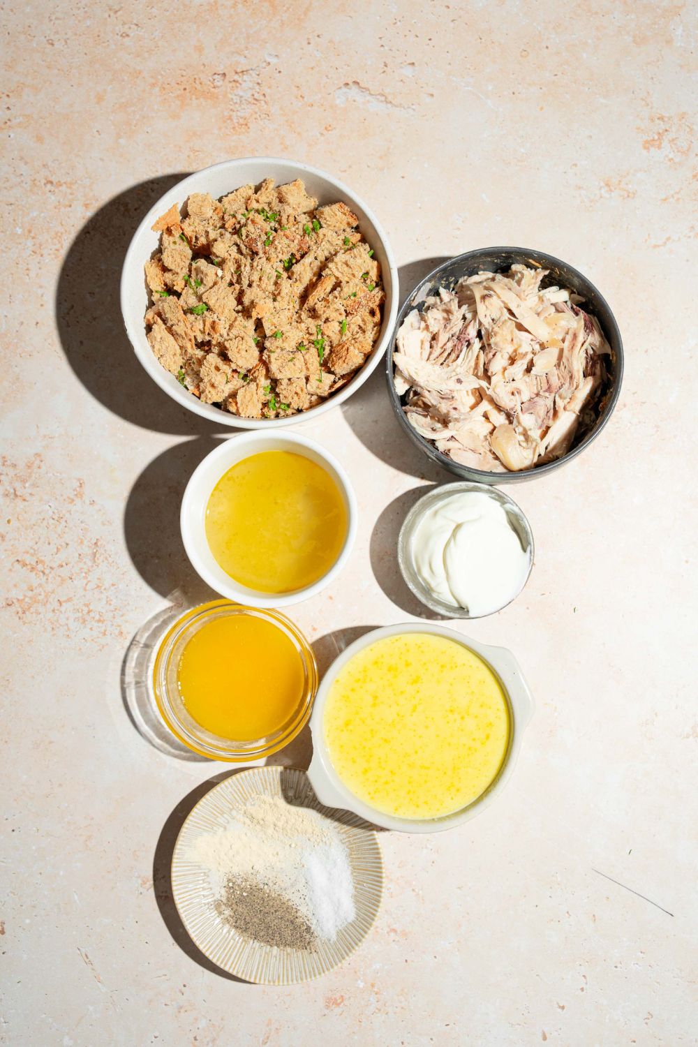 A tan counter with several bowls containing ingredients to make chicken and stuffing casserole including shredded chicken, stuffing mix, cream of chicken soup, sour cream, butter, and seasonings.
