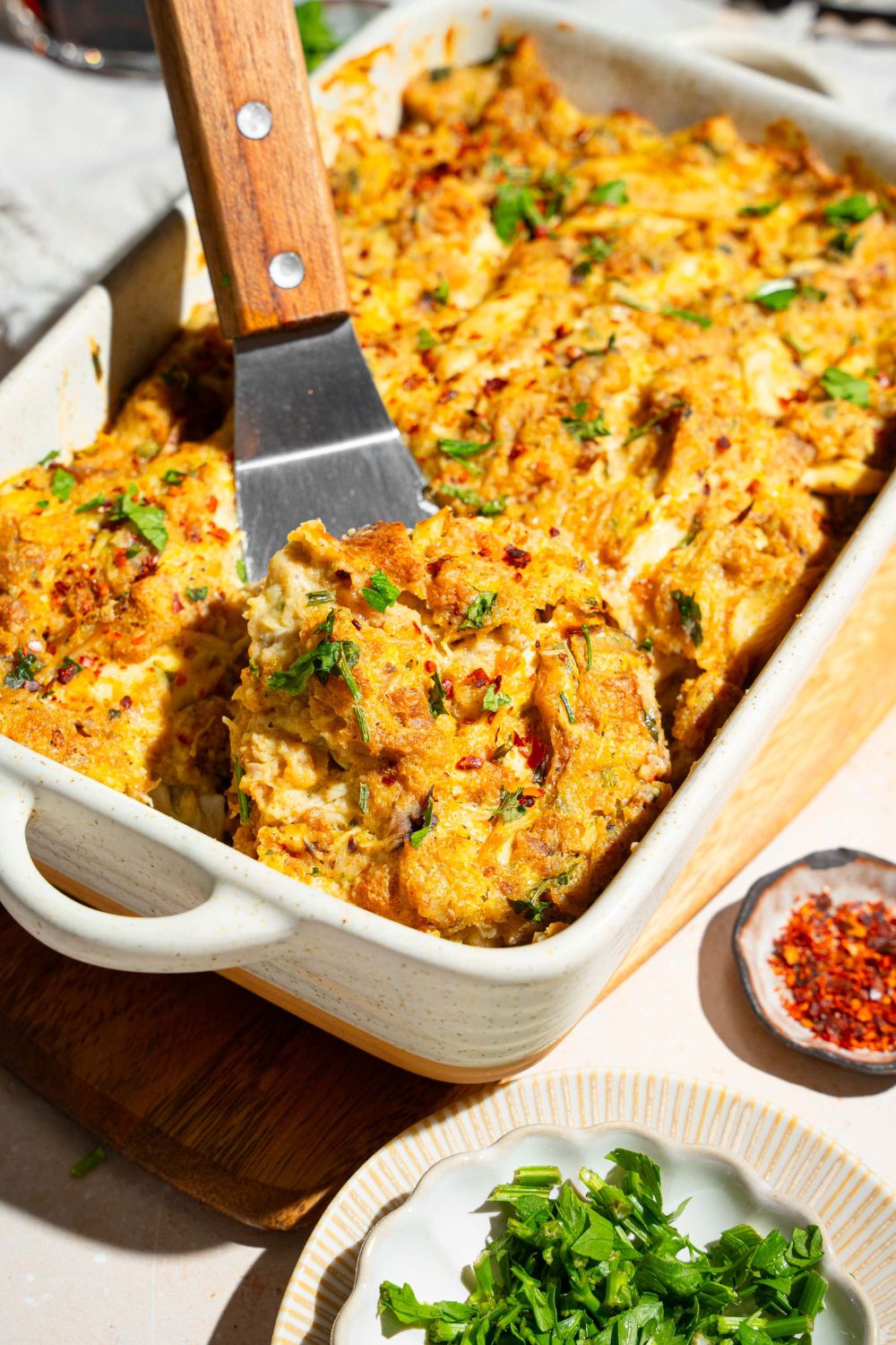 A baking dish with Dolly Parton's chicken and stuffing casserole garnished with fresh parsley. A spatula is taking a piece from the casserole.
