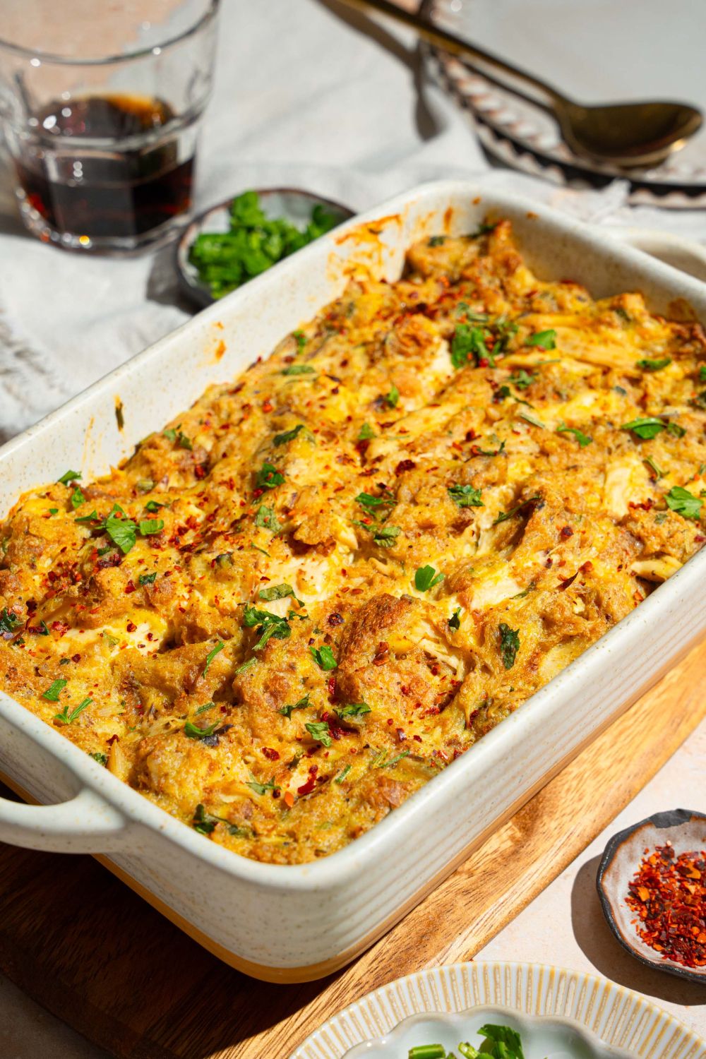 A baking dish with Dolly Parton's chicken and stuffing casserole garnished with fresh parsley. The dish s on a wooden board on a tan counter with small bowls of garnishes.
