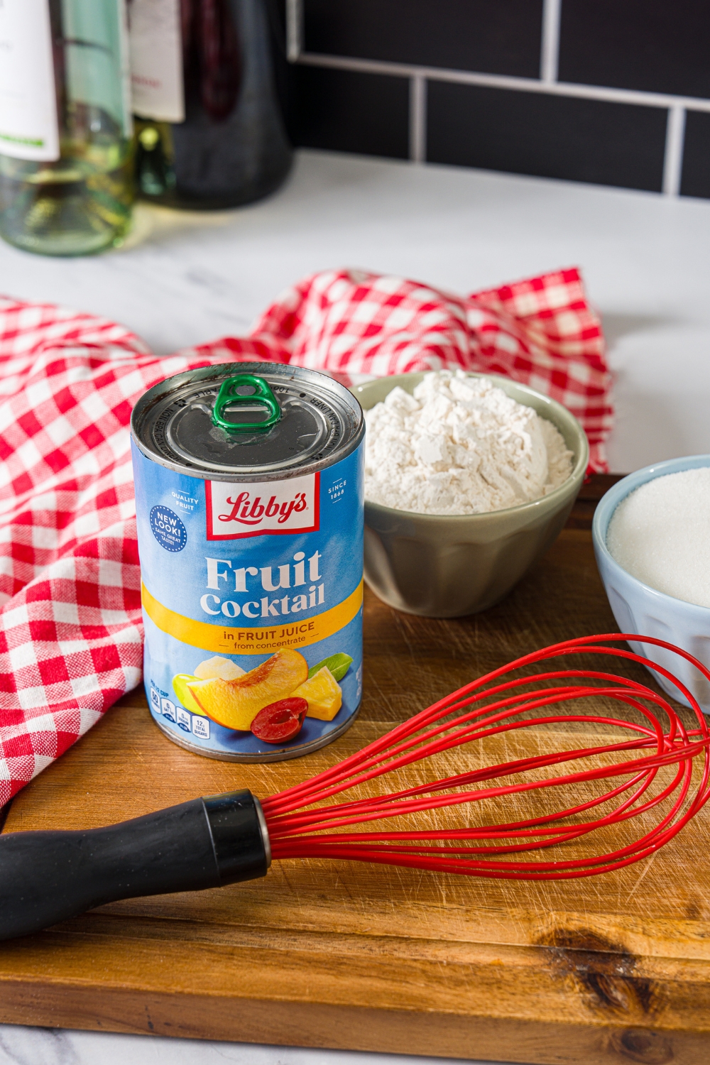 A can of fruit cocktail, a bowl of self rising flour, and a bowl of sugar with a red whisk on front of it, all on a wood cutting board.