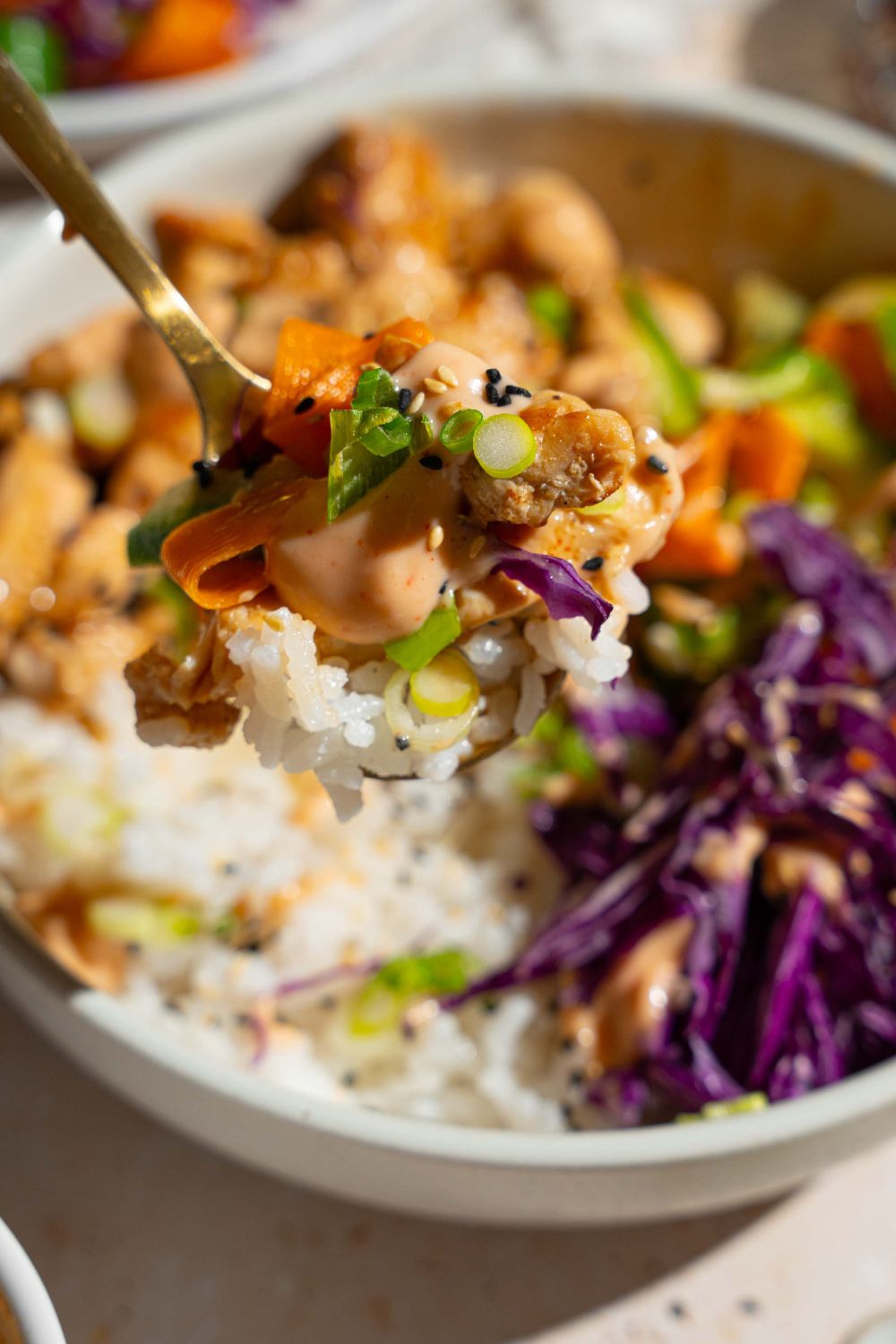 A close up of a fork with a bite of bang bang chicken bowl including rice, bang bang chicken, cabbage, cucumber carrot salad, and sesame seeds. There is a chicken rice bowl blurred in the background.