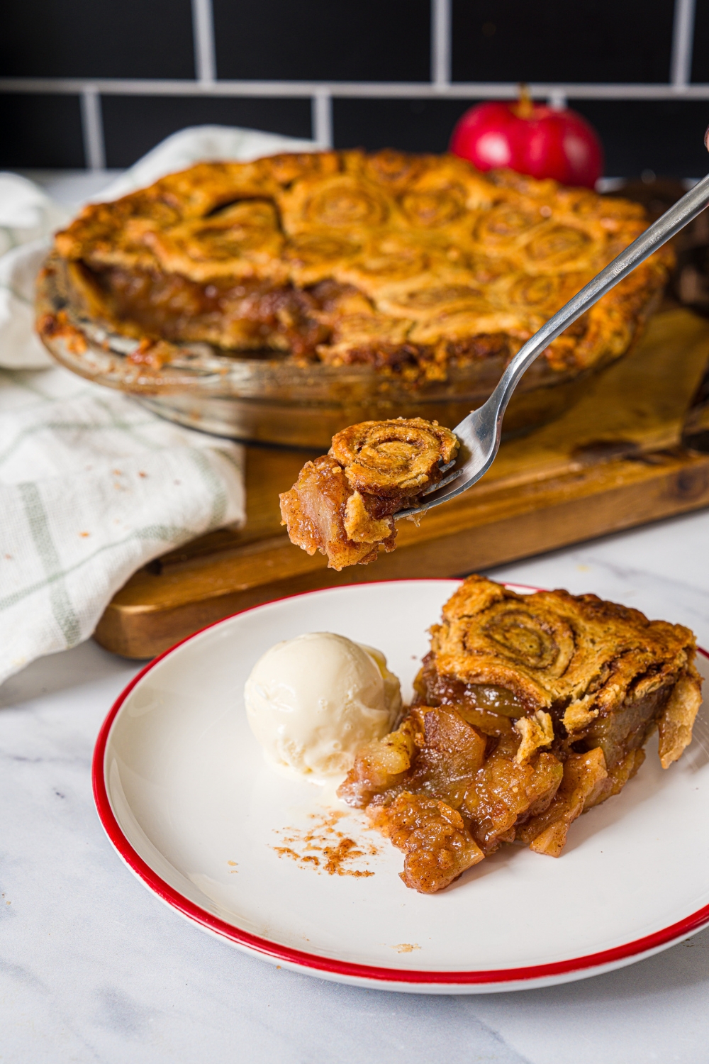 A white plate with a slice of apple pie with cinnamon roll crust served with a scoop of vanilla ice cream. A fork is taking a slice from the pie. The plate is on a marble counter.