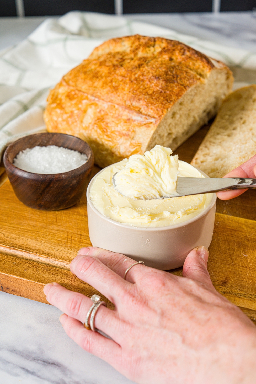 A small bowl of cultured butter. A butter knife is scooping into the butter. The bowl is on a wooden board with a bowl of salt and loaf of bread.