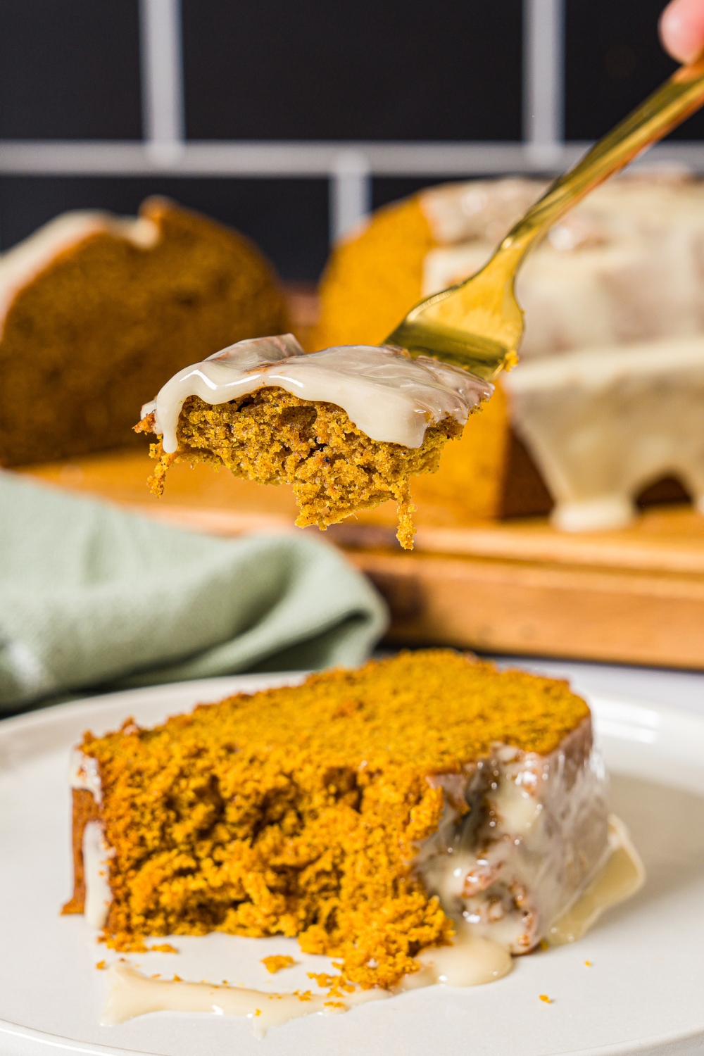 A white plate with a slice of pumpkin bread with maple glaze. There is a fork taking a bite from the slice. The plate is on a marble counter with a green cloth napkin.