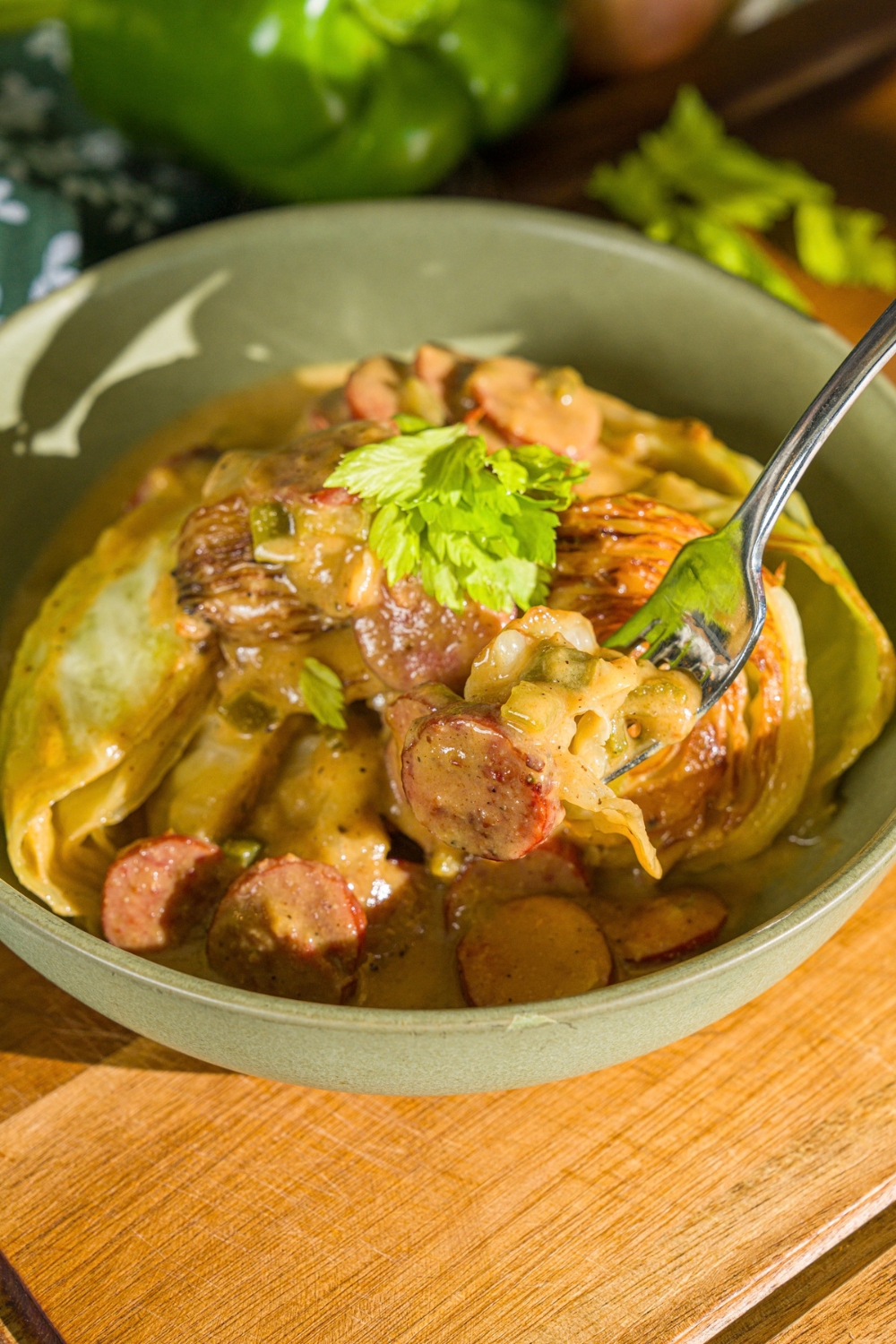 A bowl with southern smothered cabbage with sausage garnished with fresh parsley. A fork is taking a bite from the bowl. The bowl is on a wooden board.