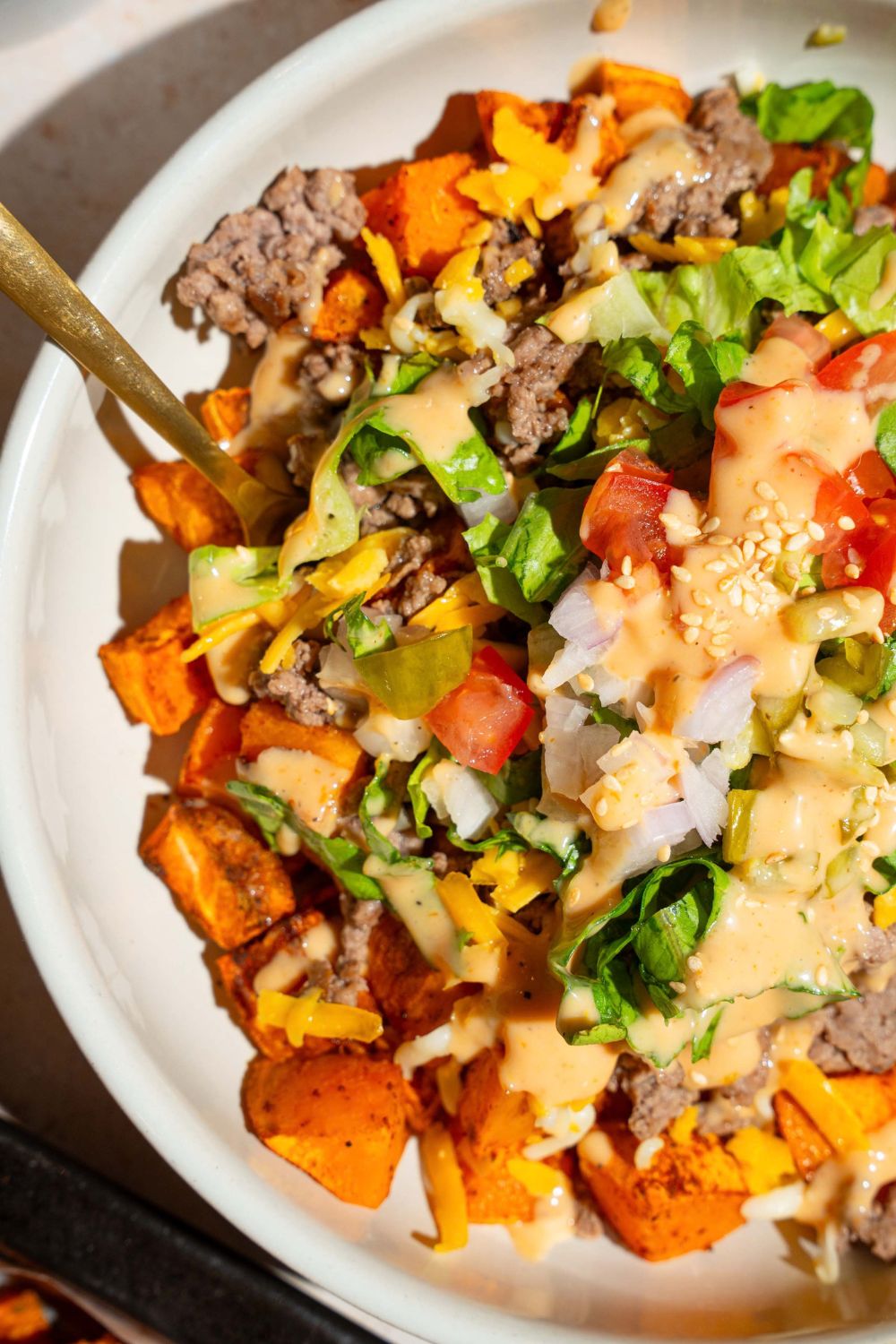 A white bowl with sweet potato burger bowls made with sweet potatoes, ground beef, shredded lettuce, tomatoes, pickles, and shredded cheese drizzled with burger sauce. There is a spoon in the bowl.