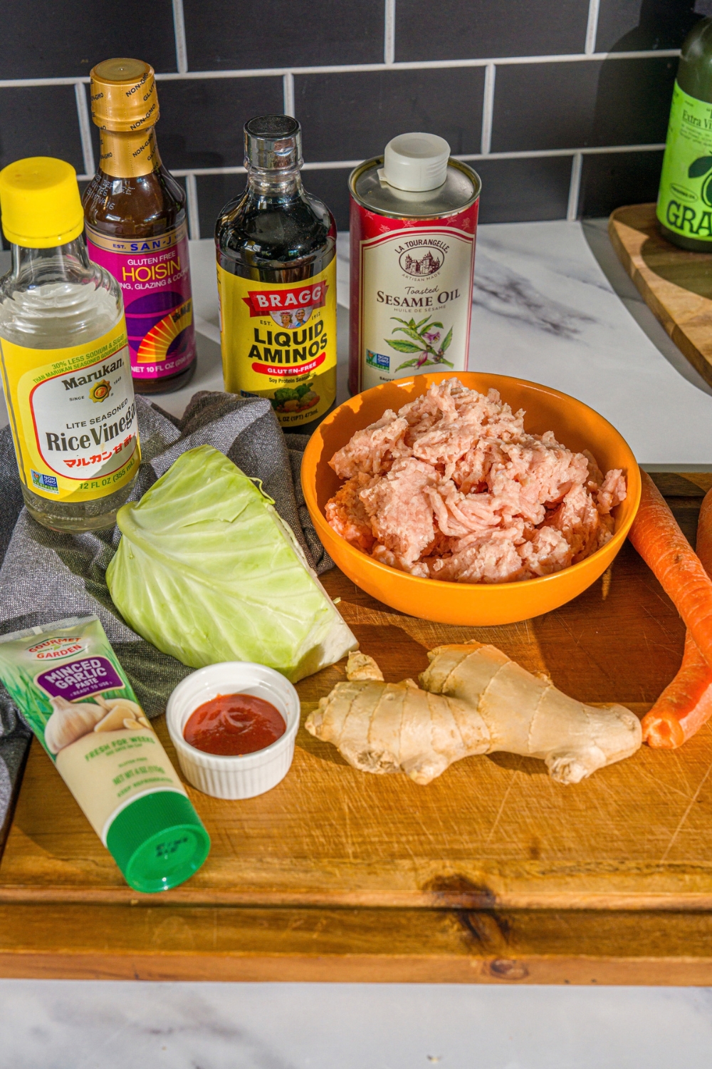 A wooden board with ingredients to make egg roll in a bowl including ground chicken, ginger, cabbage, carrot, oil, hoisin, rice vinegar, coconut aminos, and sriracha.