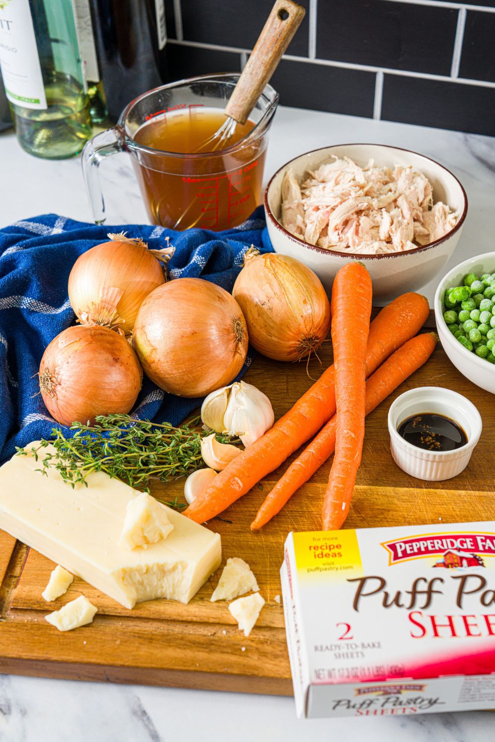 A wooden board with ingredients to make french onion chicken pot pie including carrots, onions, garlic, cheese, rosemary, peas, chicken, and more.