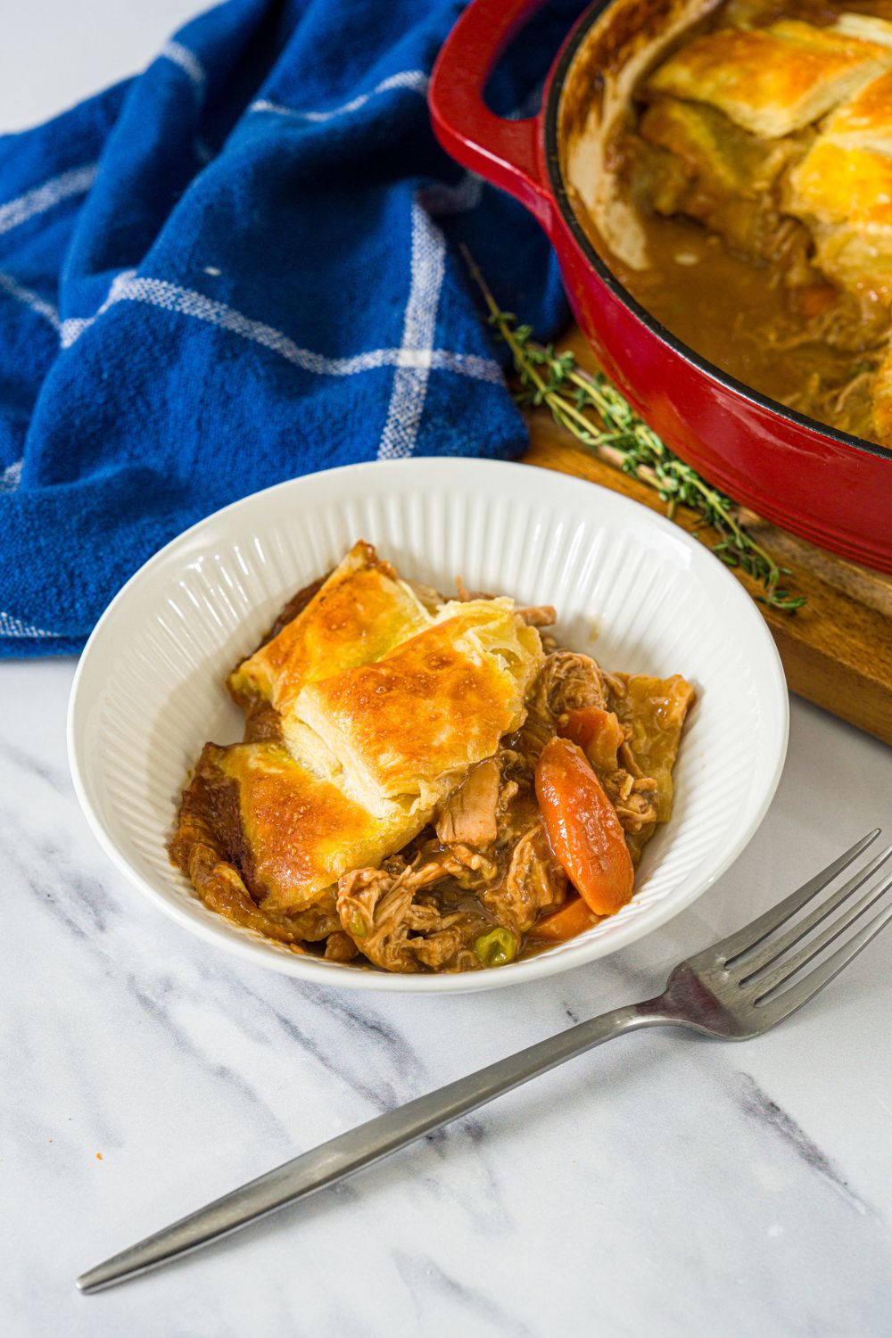 A white bowl with a slice of french onion chicken pot pie. The bowl is on a marble counter with a fork and dutch oven with pot pie.
