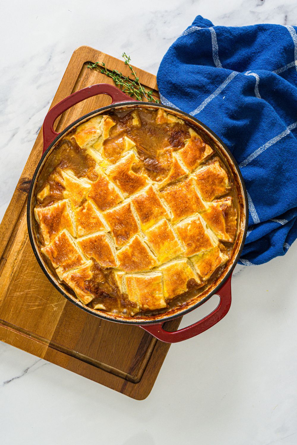 A dutch oven with baked french onion chicken pot pie. The oven is on a wooden board with a sprig of thyme and blue cloth napkin.