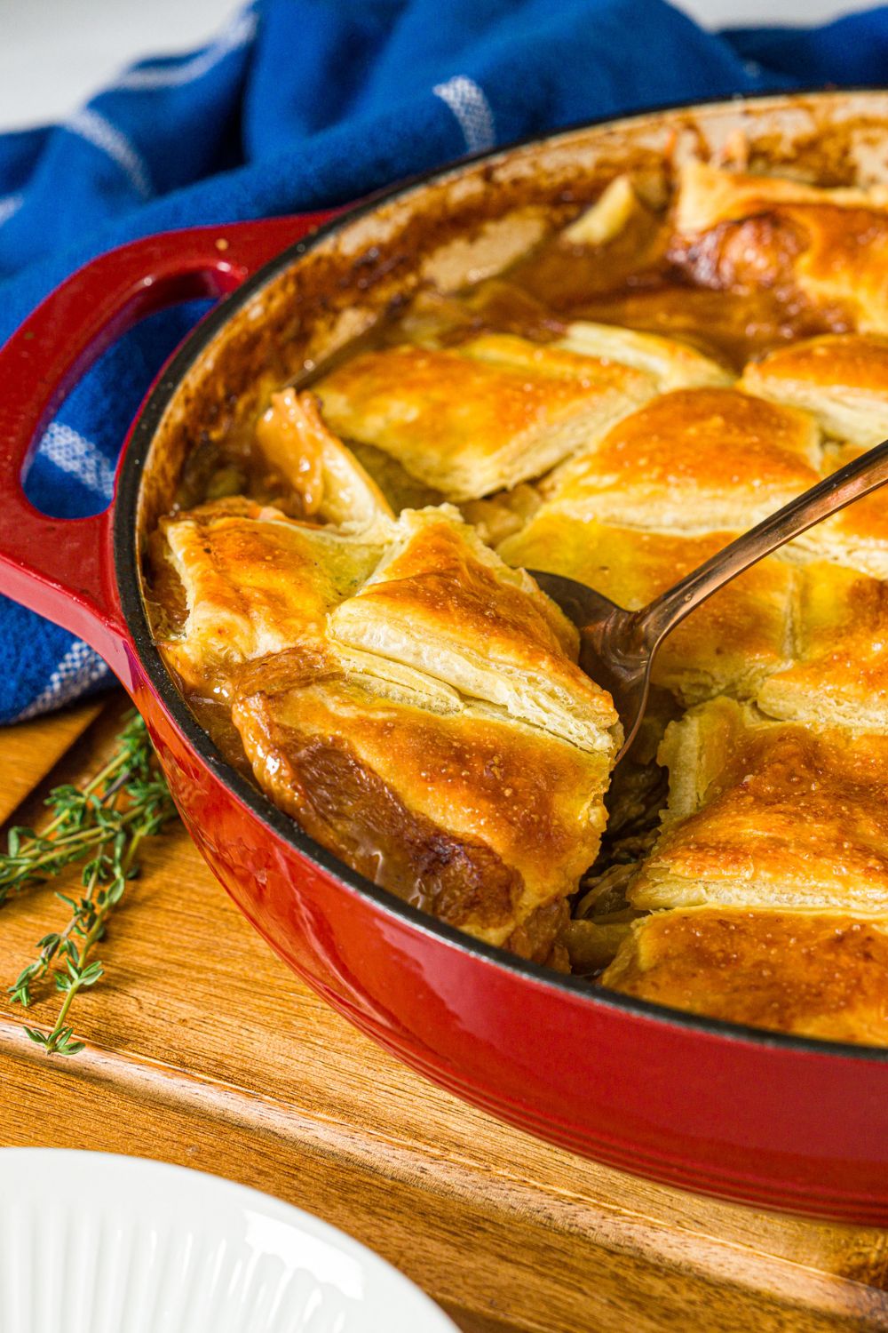 A dutch oven with baked french onion chicken pot pie. A spoon is taking a piece of pot pie from the dish. The dutch oven is on a wooden board with a sprig of thyme and blue cloth napkin.